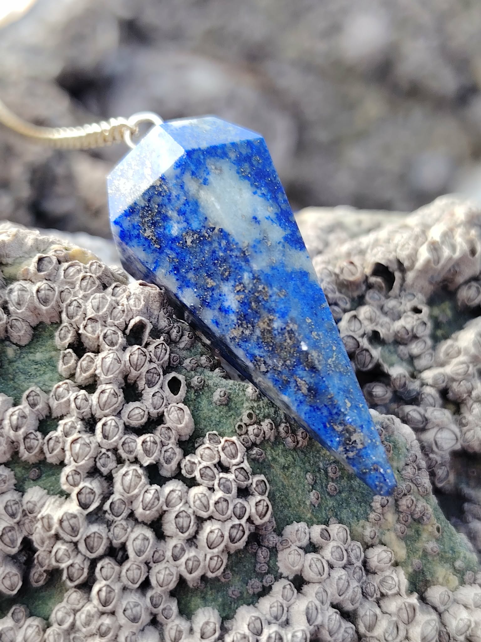 Pendule en lapis-lazuli bleu et doré posé sur un rocher marin couvert de coquillages en Bretagne, dans un décor naturel évoquant intuition, apaisement et clarté mentale.