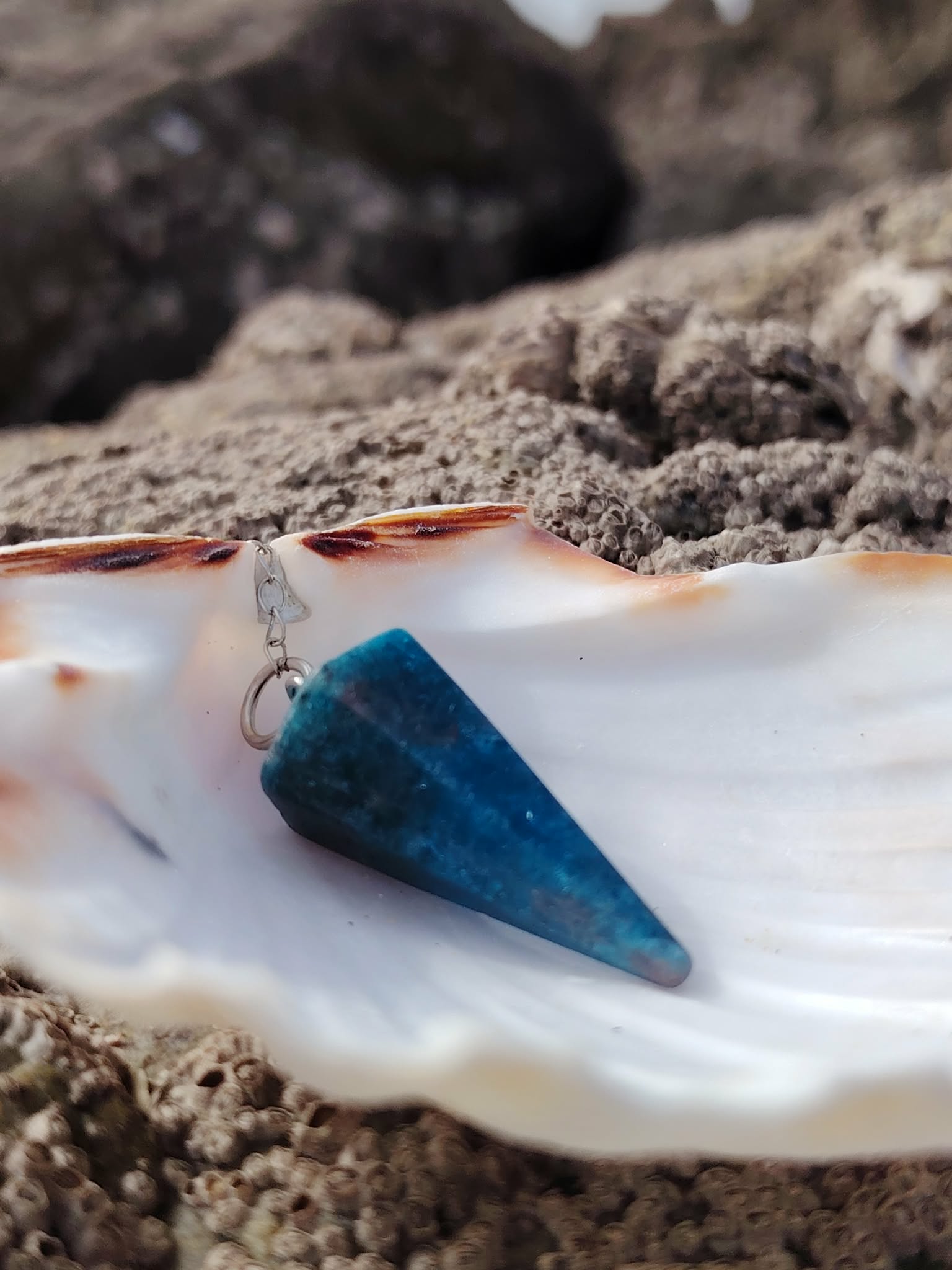 Pendule en apatite bleue posé sur un coquillage blanc, avec rocher texturé en arrière-plan, dans un décor naturel marin, évoquant intuition, apaisement émotionnel et clarté intérieure.