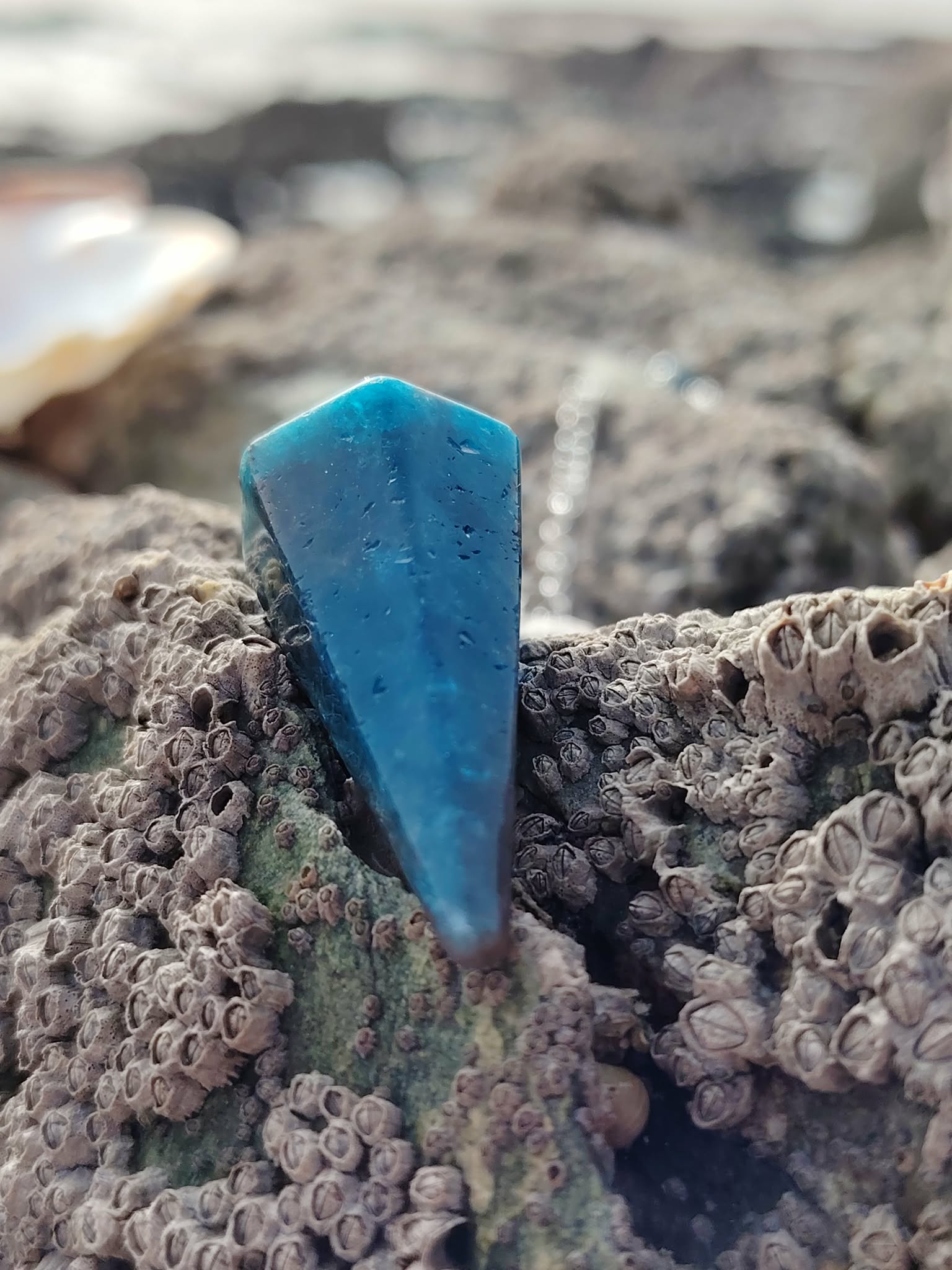 Pendule en apatite bleue posé sur un rocher breton couvert de balanes, dans un décor marin doux et minéral, photo prise en Bretagne, évoquant intuition, apaisement émotionnel et clarté intérieure.