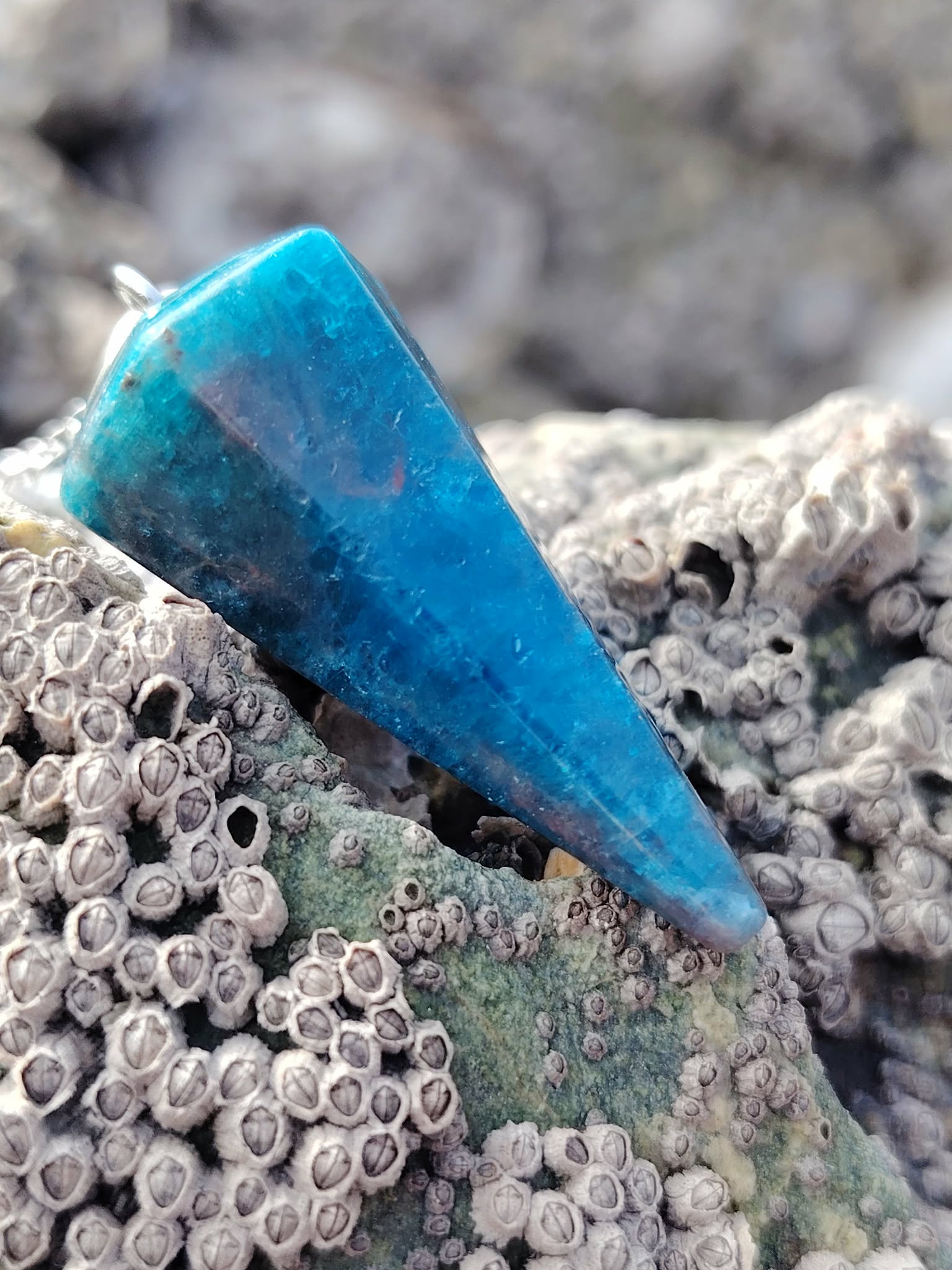 Pendule en apatite bleue posé sur un rocher couvert de balanes, en décor côtier breton aux tons minéraux, photo prise en Bretagne, évoquant intuition, apaisement émotionnel et clarté intérieure.