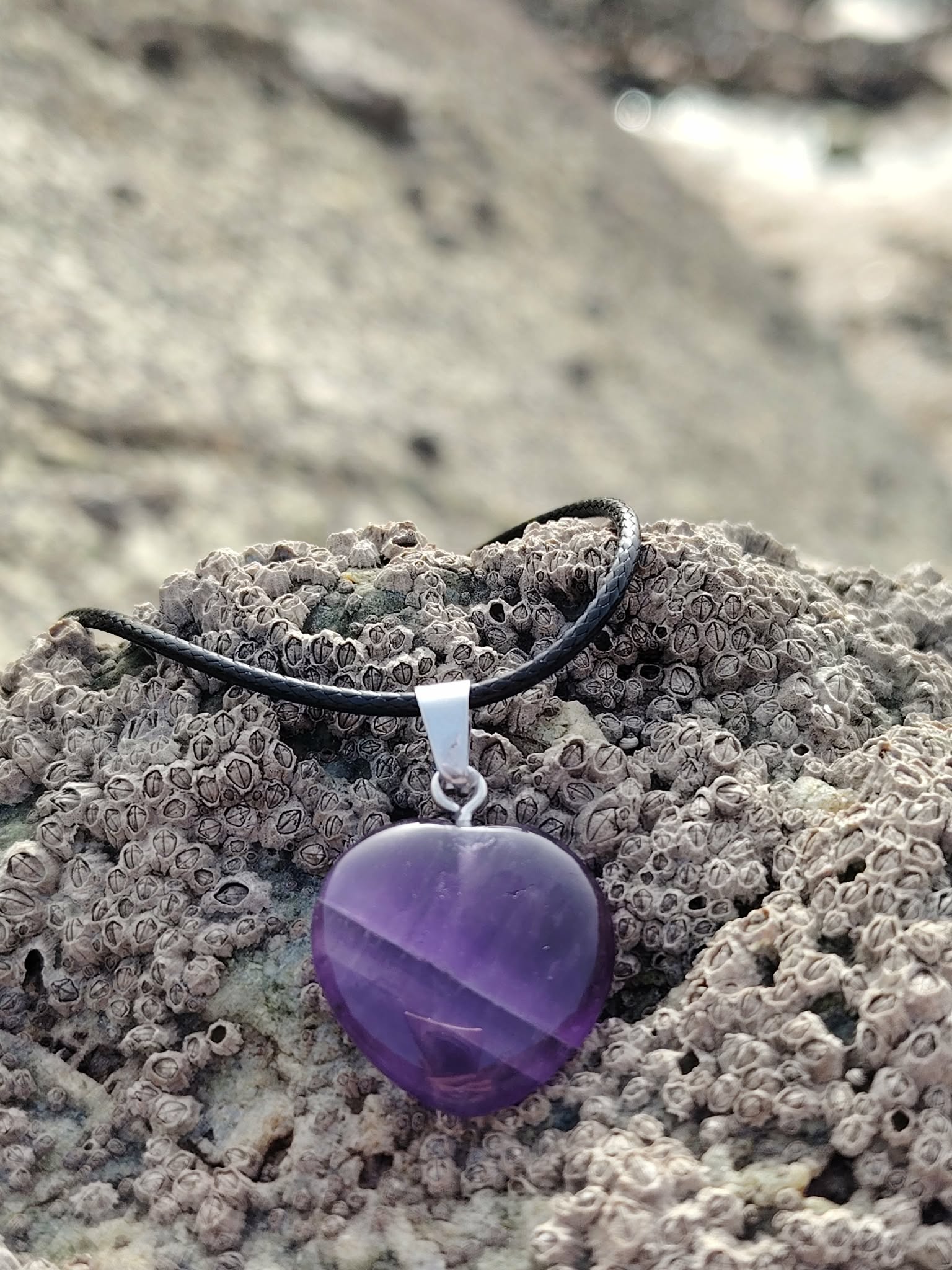 Pendentif cœur en améthyste sur cordon noir, posé sur la roche près de l’eau, dans une ambiance minérale apaisante évoquant sérénité, intuition et douceur intérieure.