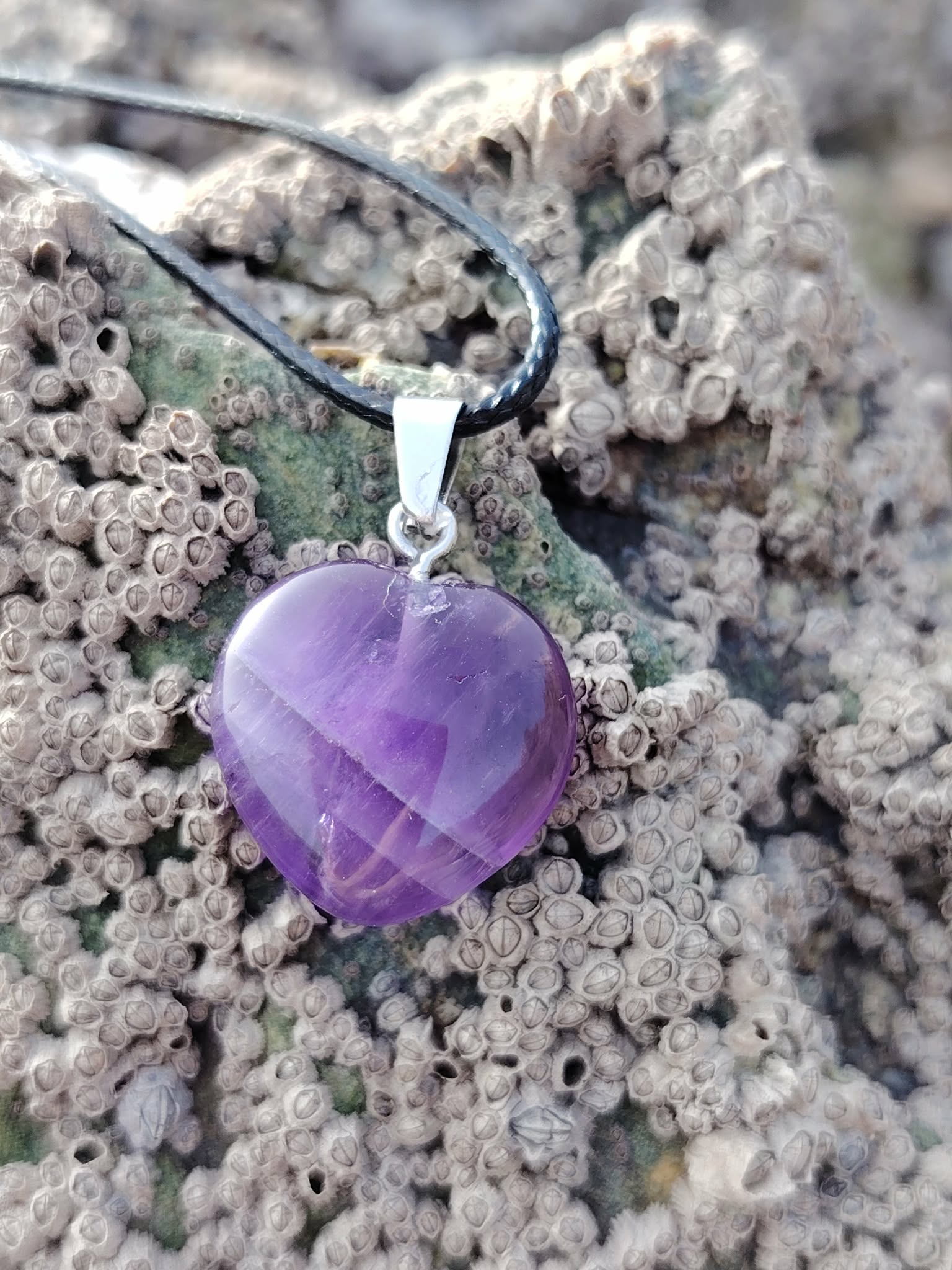 Pendentif cœur en améthyste sur cordon noir, posé sur la roche près de l’eau, dans une ambiance minérale apaisante évoquant sérénité, intuition et douceur intérieure.