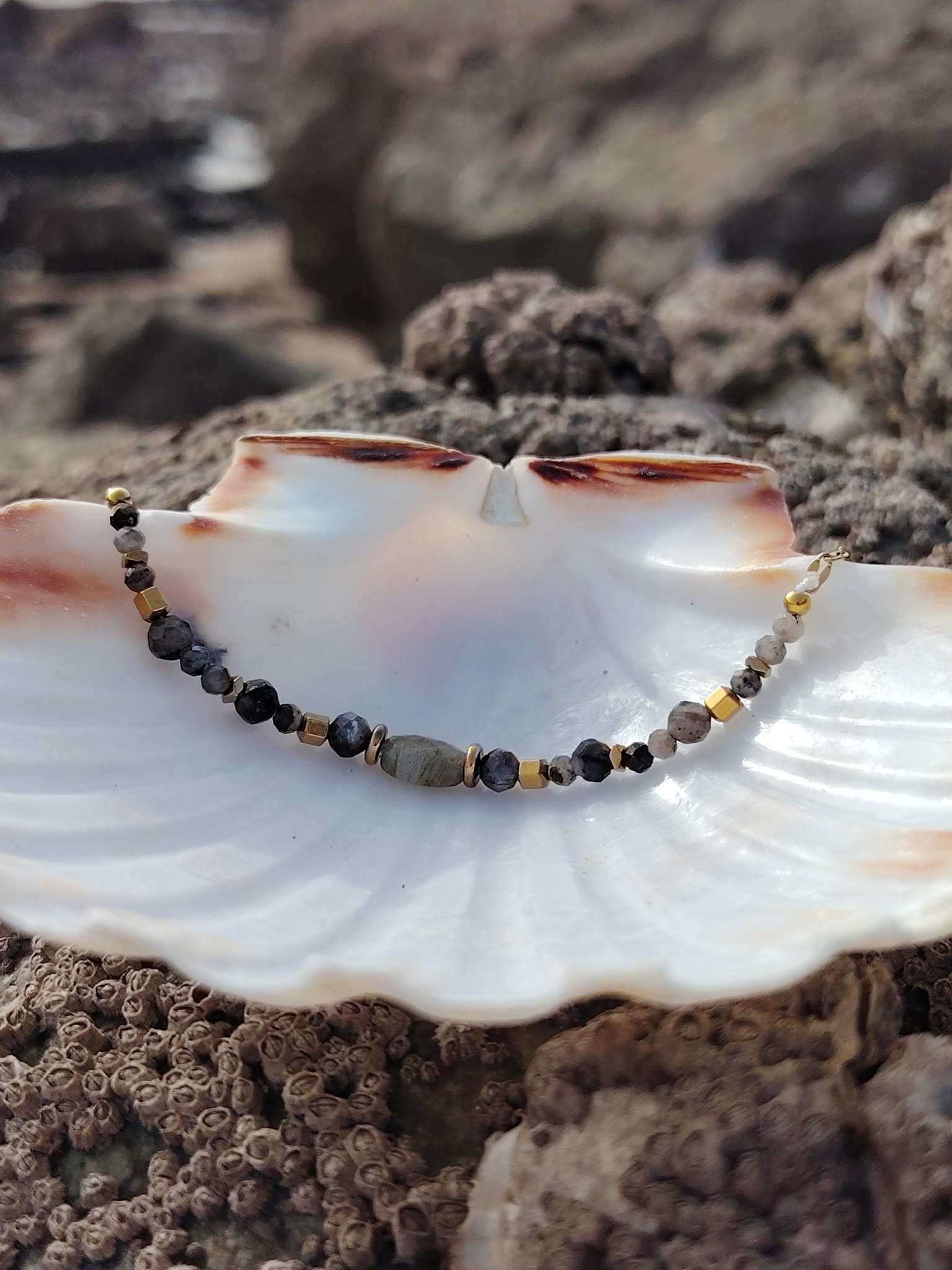 Bracelet doré en labradorite dans un coquillage blanc sur rochers bretons, reflets bleutés, apaisement et inspiration
