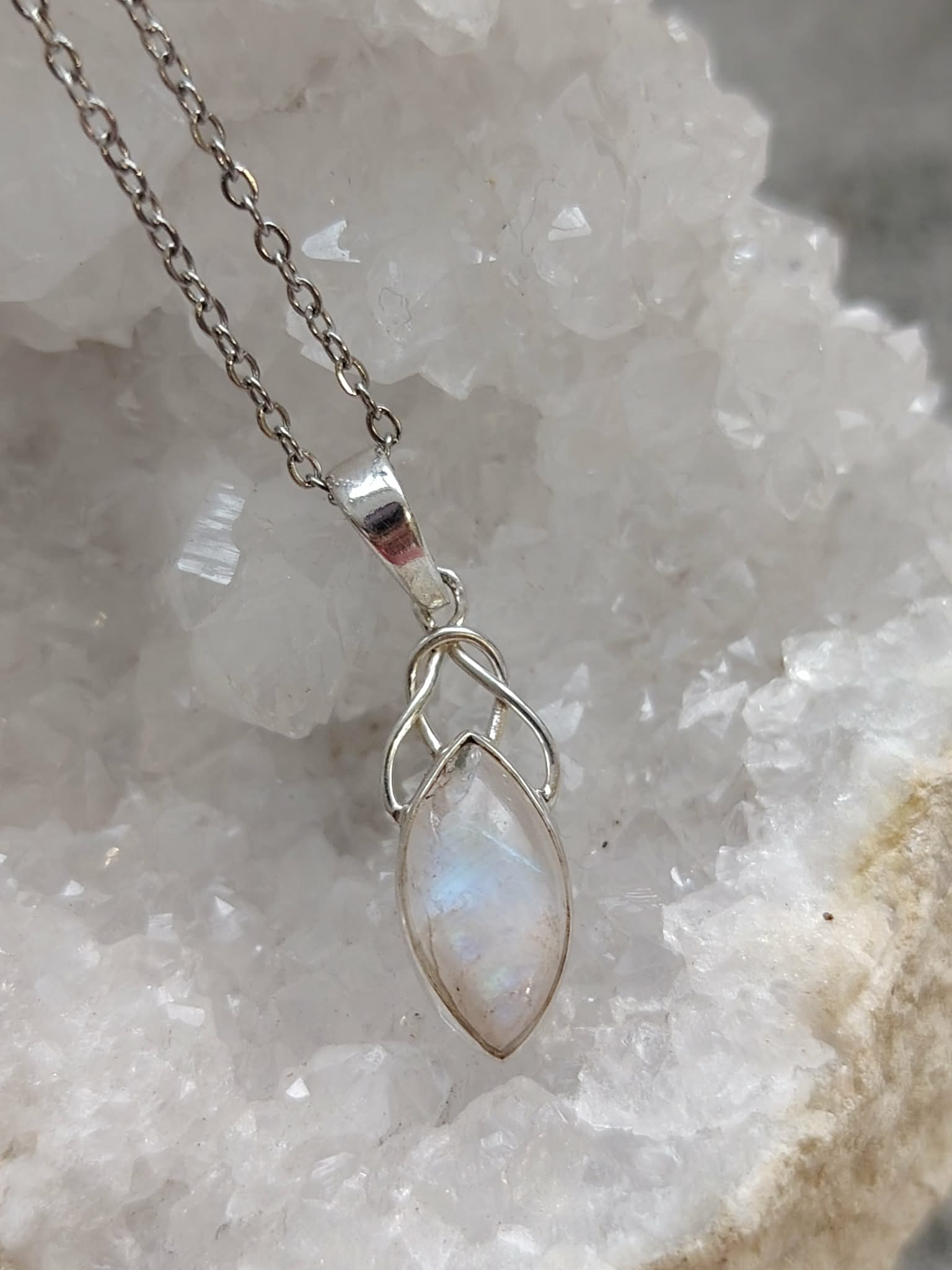 Pendentif elfique en péristérite argent 925 avec chaîne en acier inoxydable, suspendu devant un amas cristallin aux nuances douces, reflet bleuté délicat, décor minéral évoquant intuition, apaisement et sérénité.