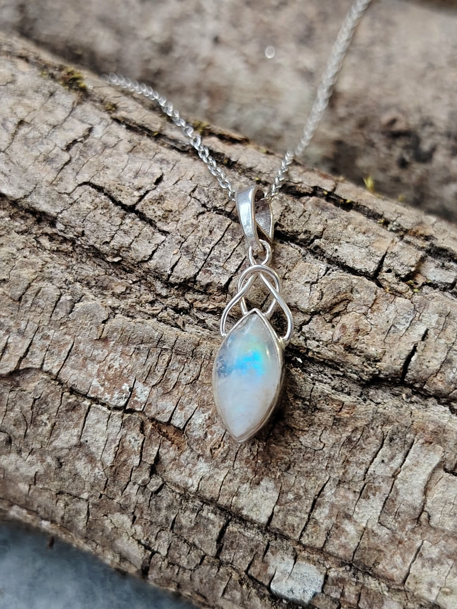 Pendentif elfique en péristérite argent 925 suspendu à une chaîne en acier inoxydable, posé sur une écorce aux reliefs bruts, reflets bleu laiteux, ambiance naturelle évoquant intuition, douceur et apaisement.