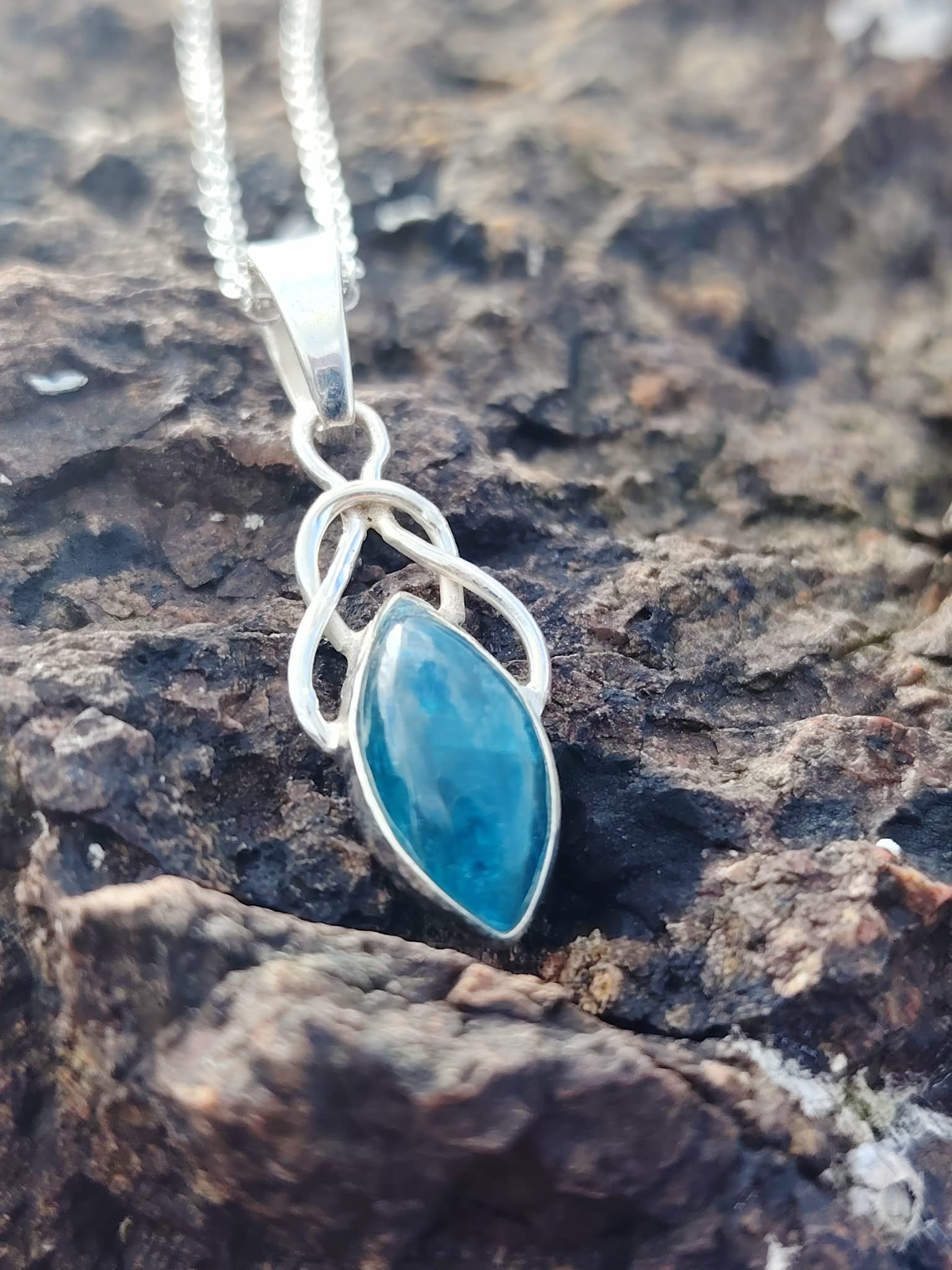 Pendentif elfique en apatite bleue monté en argent 925, photographié sur des rochers bretons sombres, collier en pierre naturelle associé à la créativité, à la confiance douce et à la paix émotionnelle.