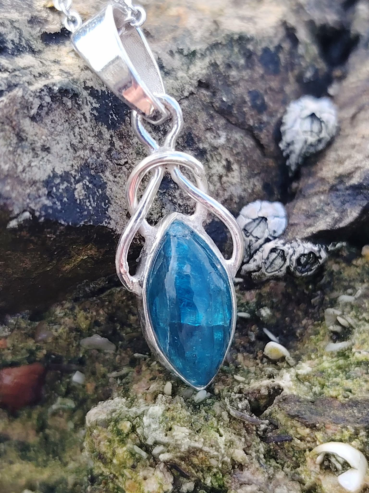 Pendentif elfique en apatite bleue et argent 925 photographié sur des rochers bretons au bord de l’eau, bijou en pierre naturelle évoquant la lucidité, l’expression sincère et l’apaisement.