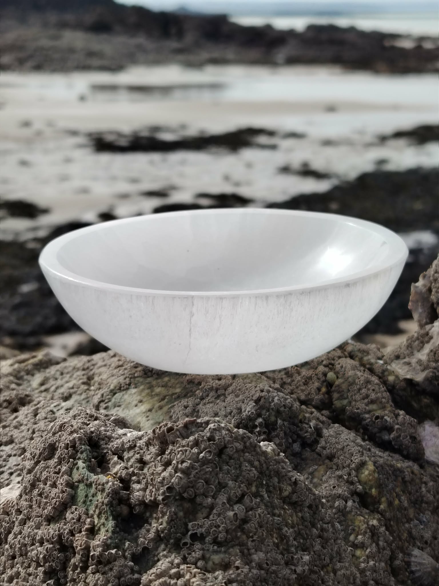 Bol en sélénite 15 cm posé sur des rochers en Bretagne face à la mer, dans une ambiance lumineuse et apaisante, inspirant purification, calme et clarté intérieure