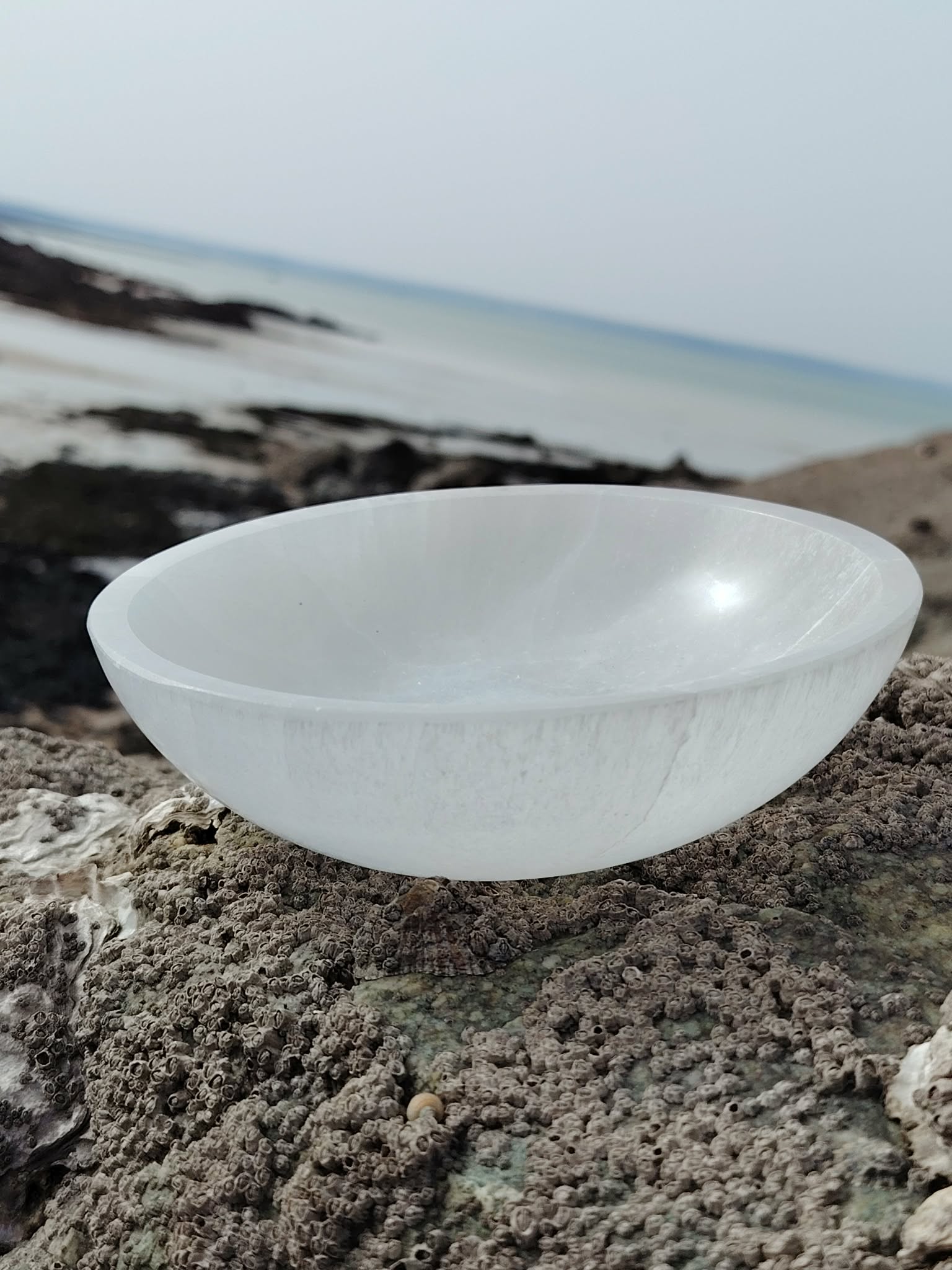 Bol en sélénite 15 cm posé sur un rocher en Bretagne face à la mer, sous une lumière douce et un ciel clair, évoquant apaisement, purification et clarté intérieure.
