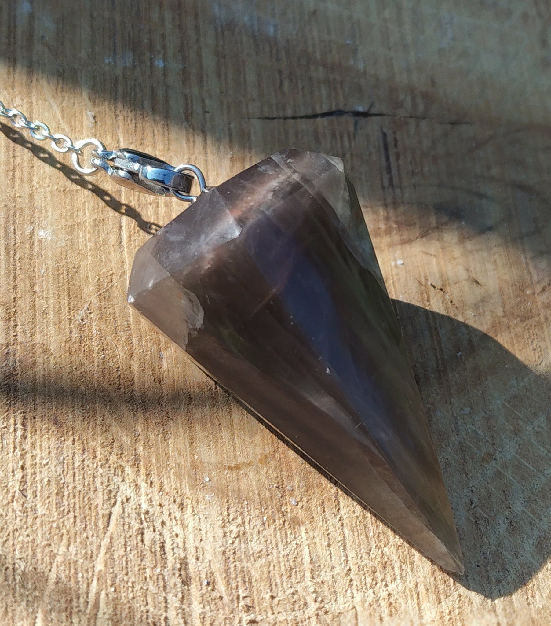 Pendule en pierre de lune noire posé sur une surface en bois clair, éclairé par une lumière chaude, avec chaîne argentée, dans une ambiance sobre et mystique évoquant intuition, ancrage et protection intérieure.