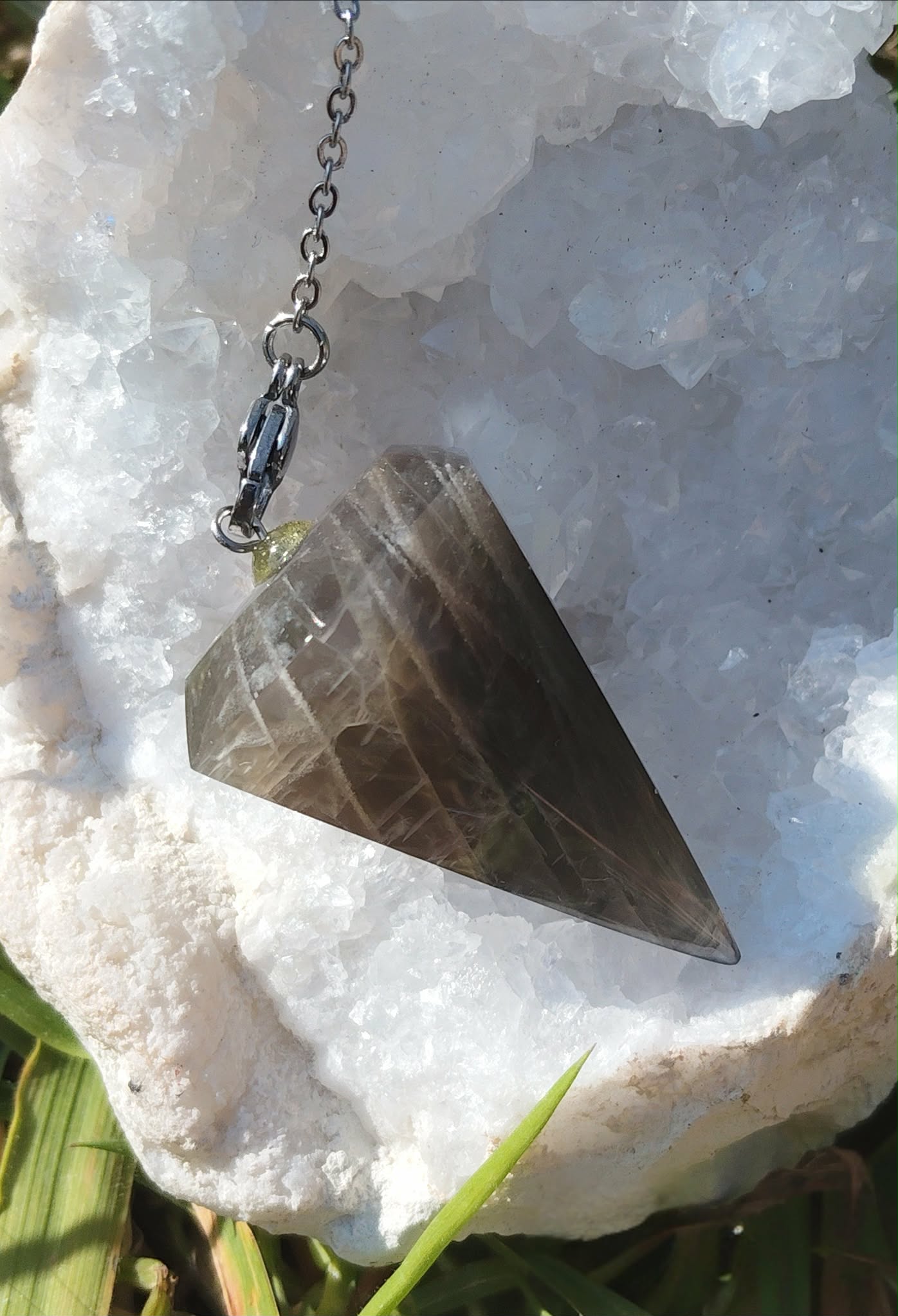 Pendule en pierre de lune noire sur un amas de quartz , baigné de lumière naturelle, dans une ambiance douce et épurée évoquant intuition, ancrage et protection intérieure.