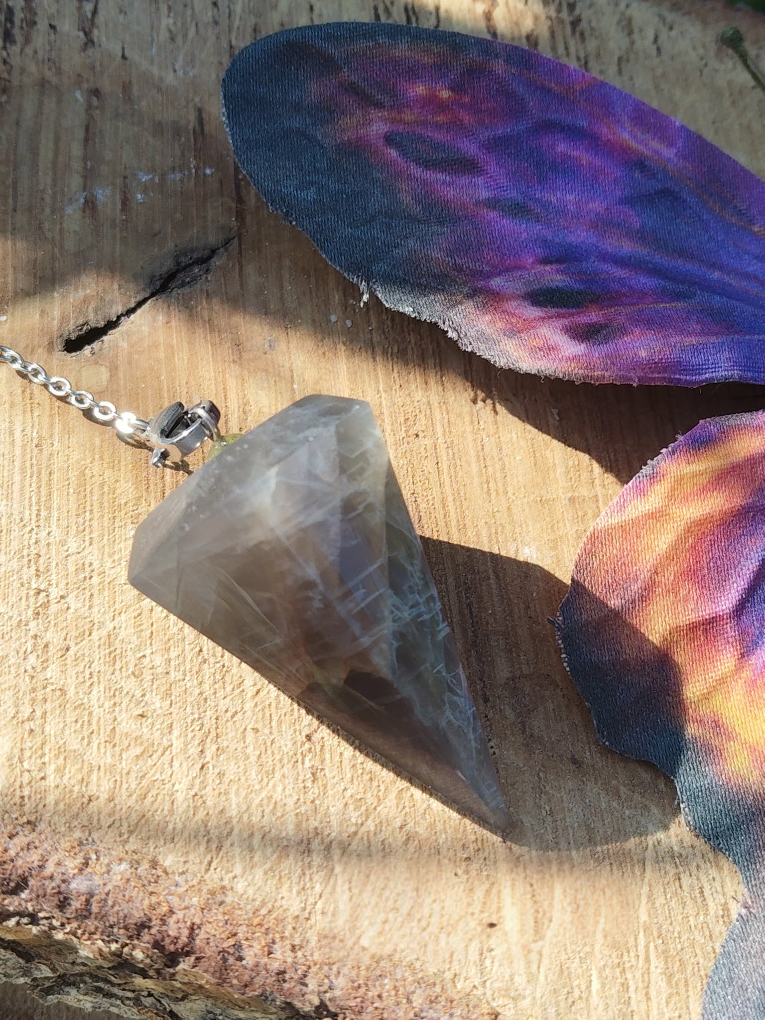 Pendule en pierre de lune noire posé sur une surface en bois clair, près de fleurs violettes et pêche, dans une ambiance lumineuse et mystique évoquant intuition, ancrage et protection intérieure.