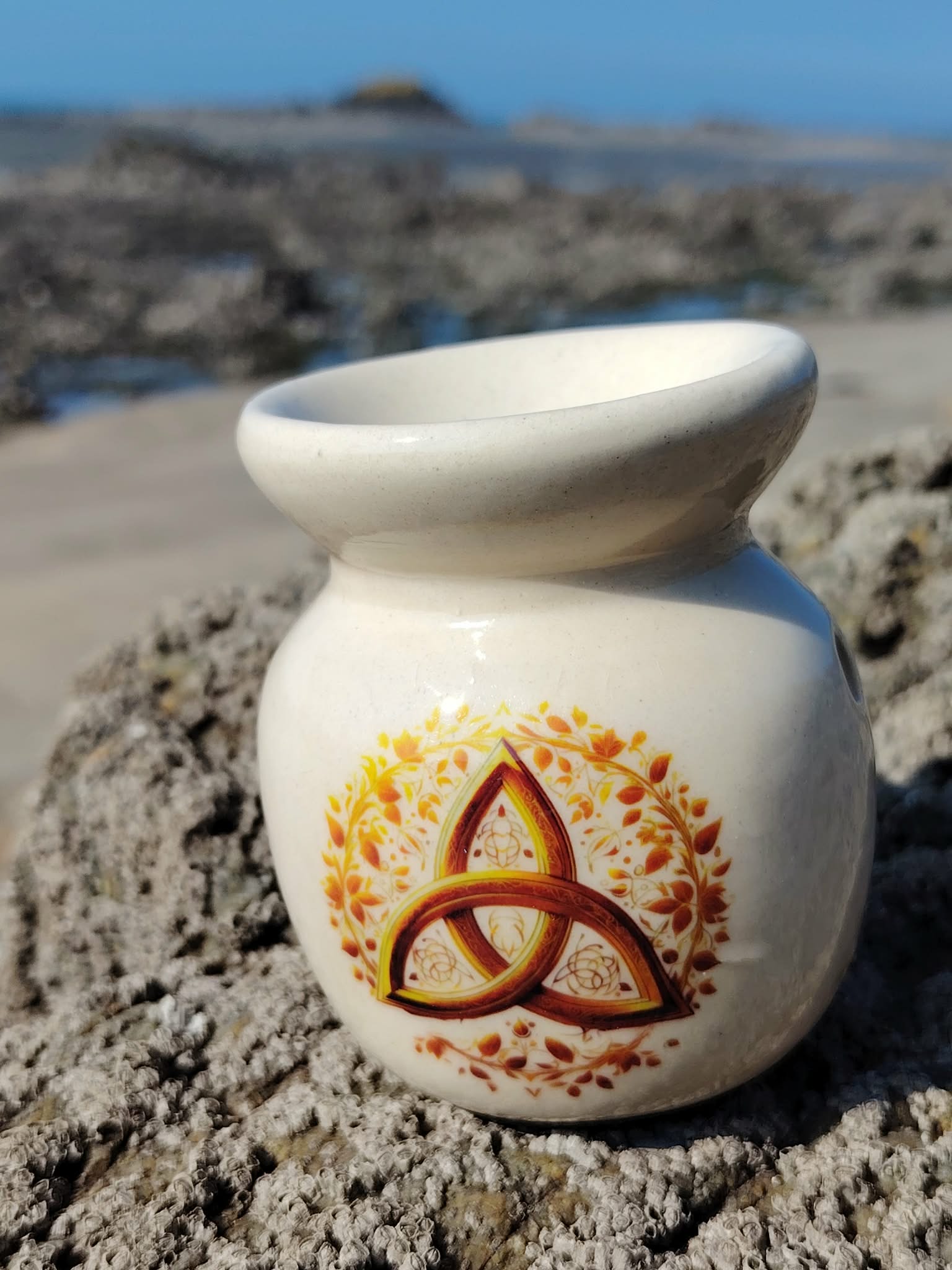 Brûleur en céramique blanc avec triquetra dorée, posé sur les rochers en Bretagne face à la mer, idéal pour fondants parfumés et bougie chauffe-plat, dans une ambiance d’harmonie et de douceur.