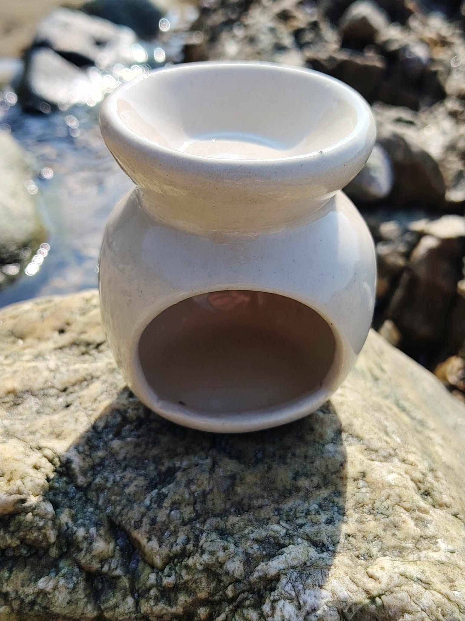 Vue arrière d’un brûleur en céramique blanc posé sur les rochers en Bretagne, avec ouverture pour bougie chauffe-plat et coupelle pour fondants, dans une ambiance douce et apaisante.