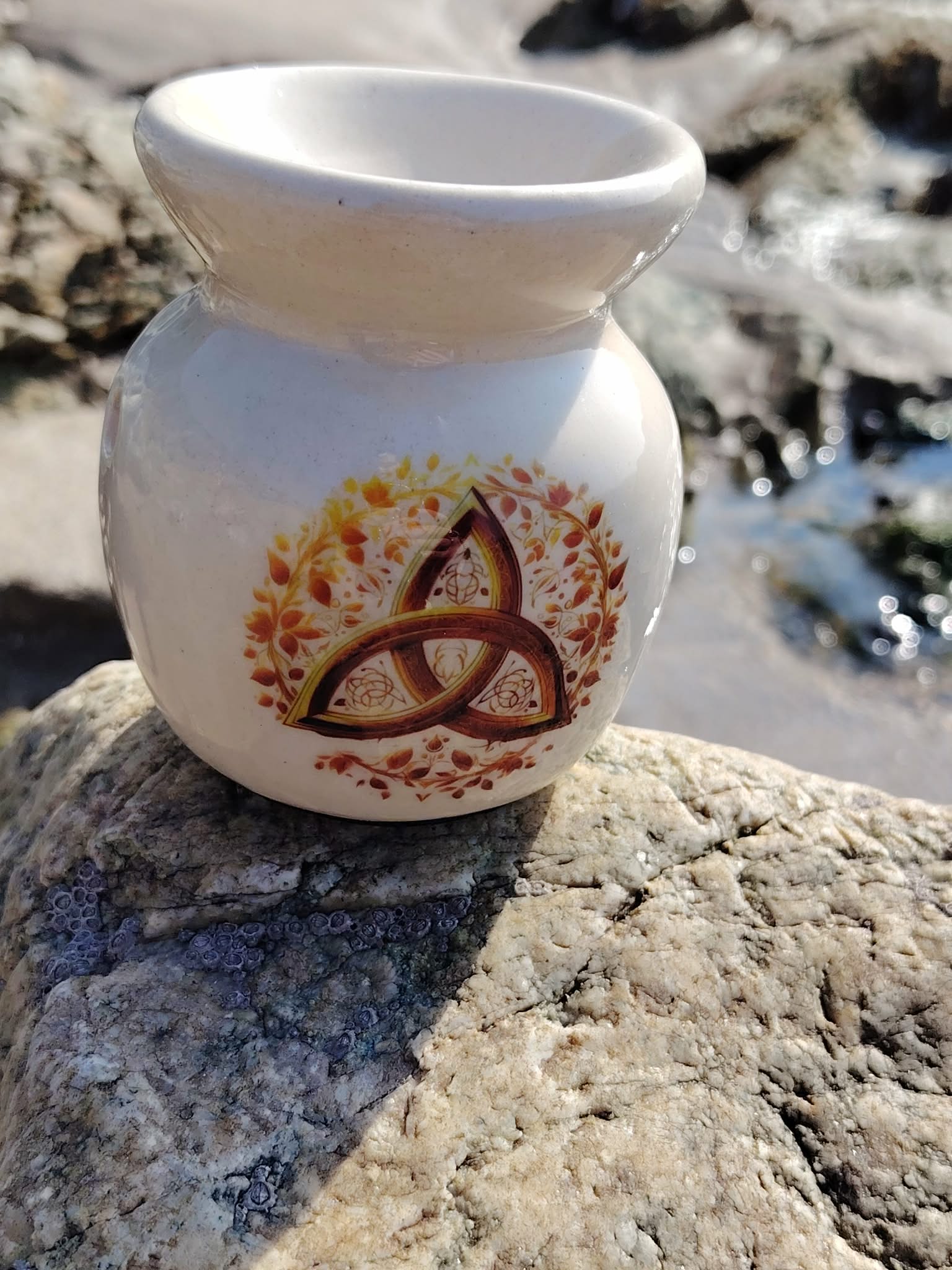 Brûleur en céramique blanc avec triquetra dorée, posé sur les rochers bretons près de l’eau, pour fondants parfumés et bougie chauffe-plat, dans une ambiance sereine et harmonieuse.