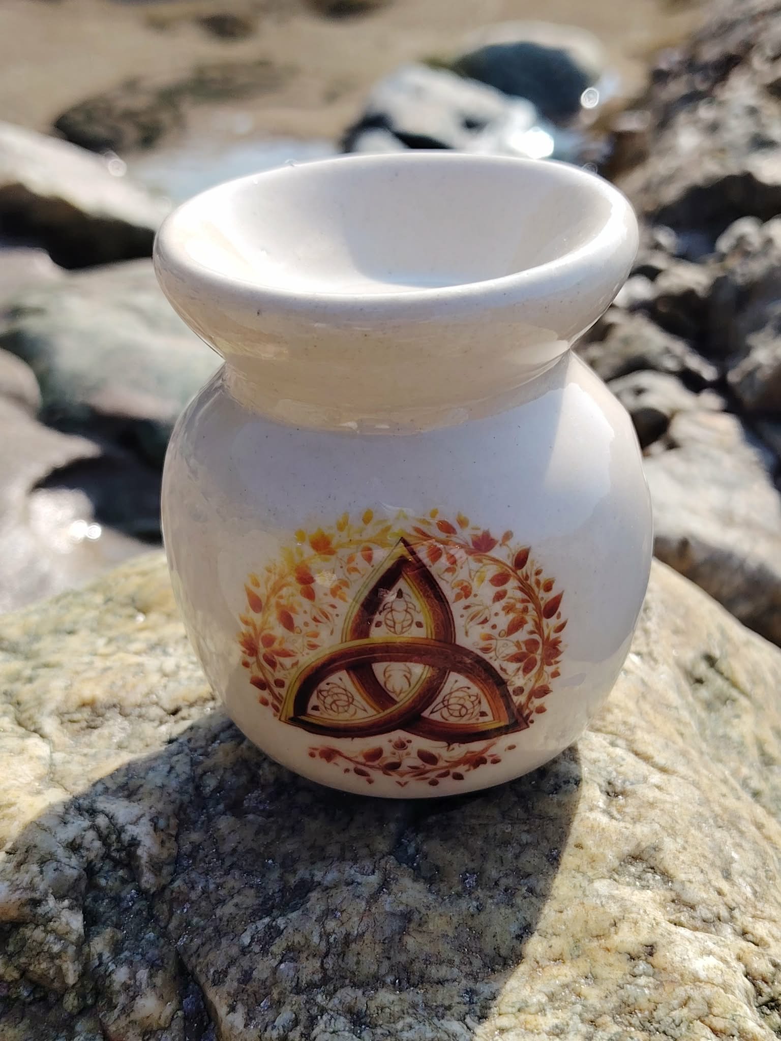 Brûleur en céramique blanc décoré d’une triquetra, posé sur un rocher en Bretagne parmi les pierres claires, dans une atmosphère douce de calme, d’harmonie et de recentrage.