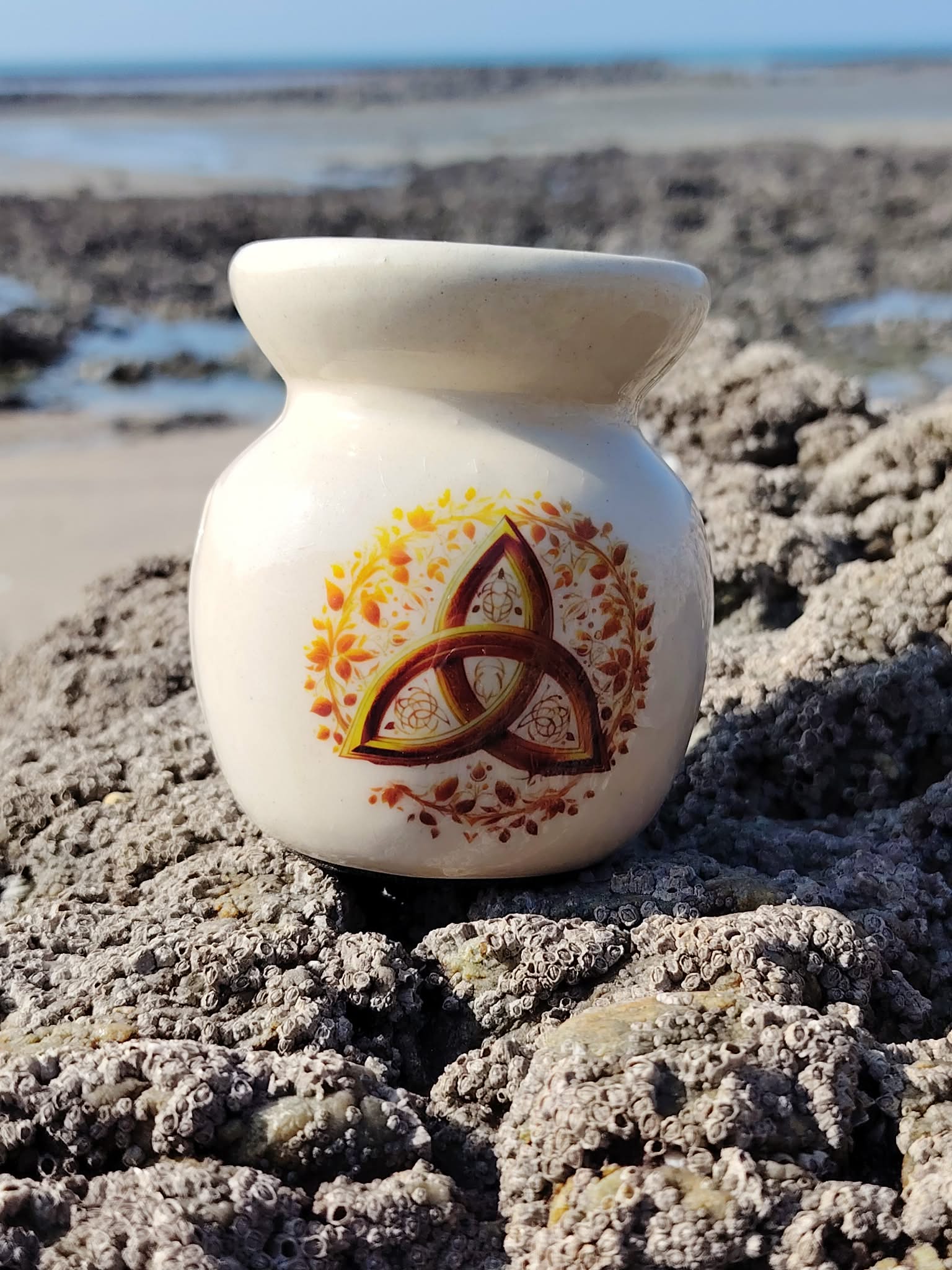 Brûleur en céramique blanc orné d’une triquetra dorée, posé sur les rochers en Bretagne face à la mer, dans une lumière douce, évoquant apaisement, harmonie et ancrage