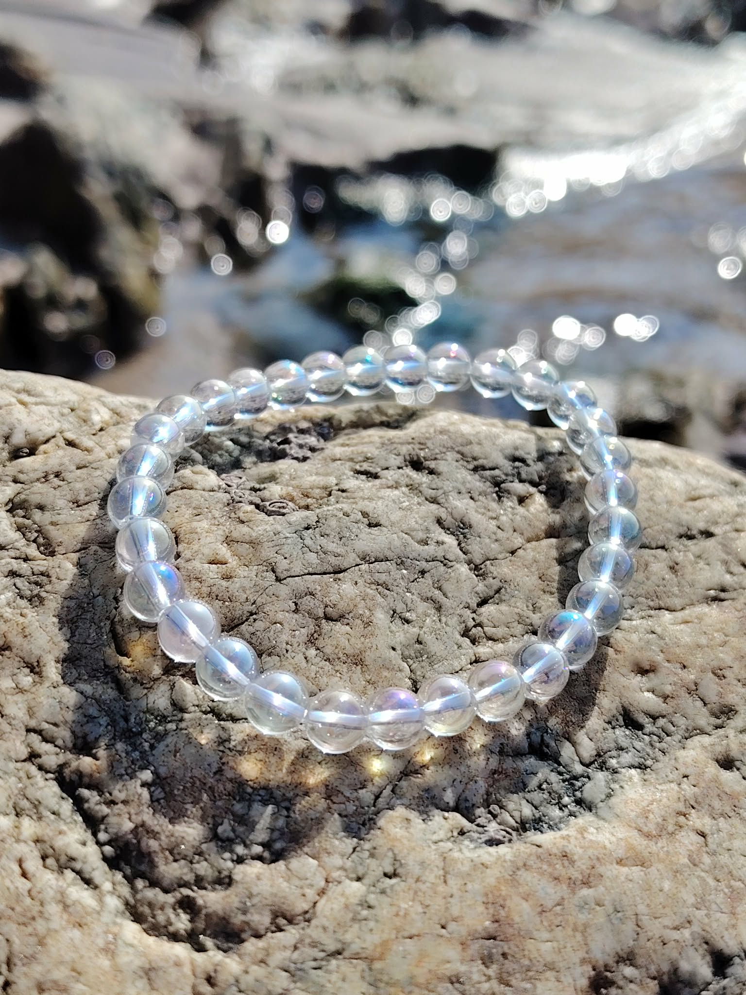 Bracelet Angel Aura Quartz 6 mm face à la mer sur rochers bretons, reflets irisés bleutés, intuition et sérénité