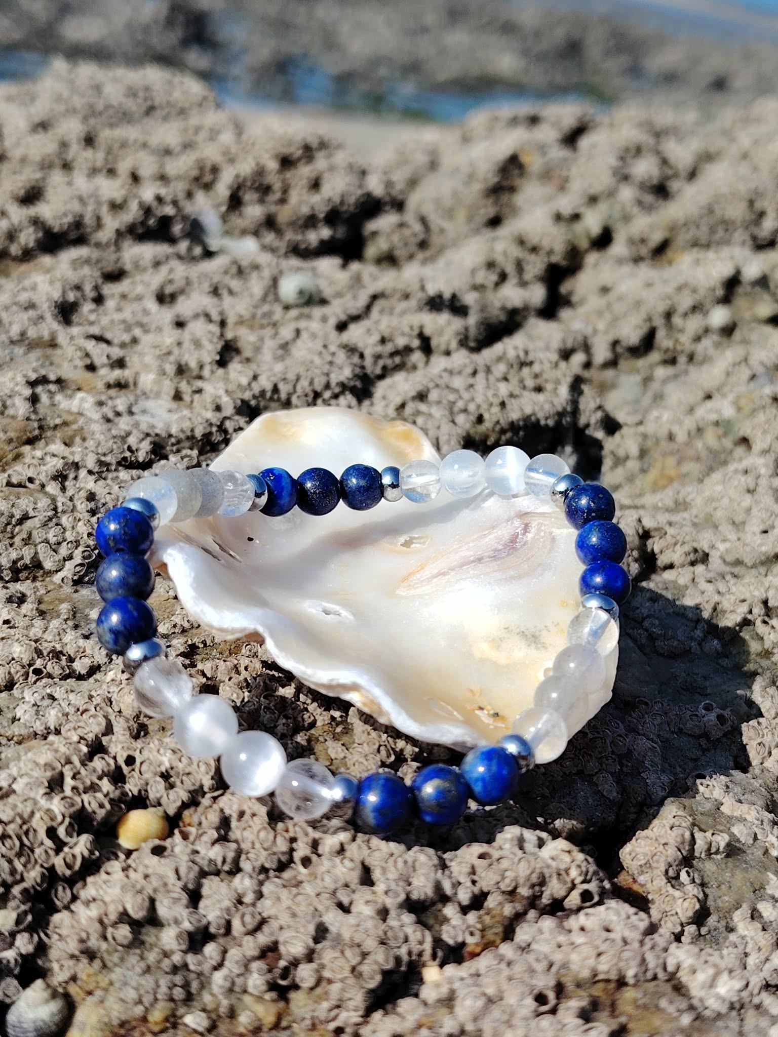 Bracelet migraine 6 mm en lapis-lazuli, labradorite et sélénite dans un coquillage sur rochers bretons face à la mer, apaisement du mental et clarté intérieure