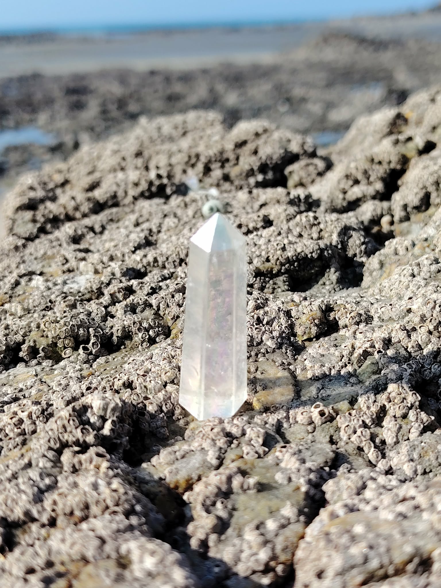 Pointe Angel Aura posée sur les rochers en Bretagne face à l’horizon, cristal irisé évoquant l’ouverture, la douceur émotionnelle et la tranquillité du mental.
