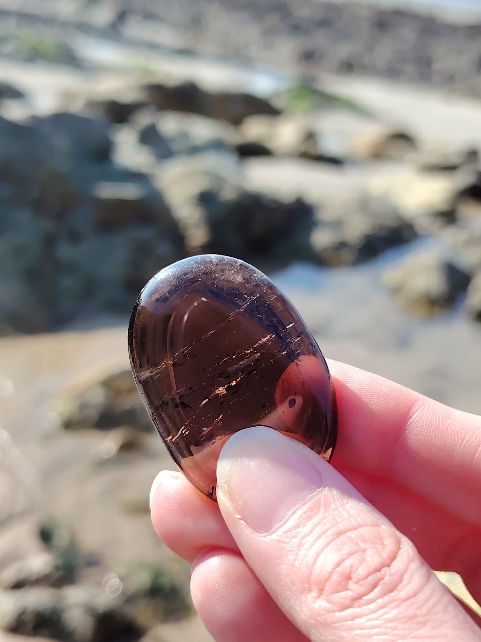 Main tenant un galet en quartz fumé face aux rochers et à l’eau en Bretagne, pierre naturelle évoquant la stabilité, le relâchement intérieur et la présence à soi.