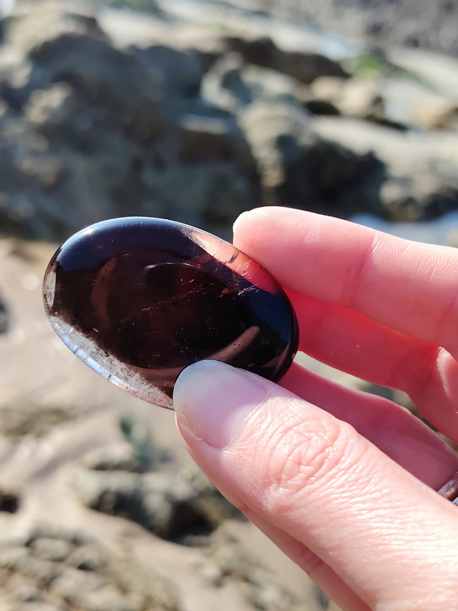 Main tenant un galet en quartz fumé devant les rochers bretons, pierre naturelle associée au discernement, à la stabilité émotionnelle et au retour au calme.