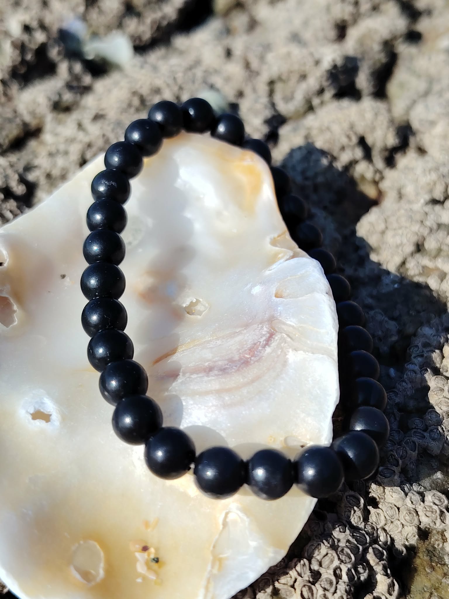 Bracelet shungite 6 mm posé sur coquillage clair avec roche texturée en fond, lucidité sobre, apaisement nerveux et protection rassurante