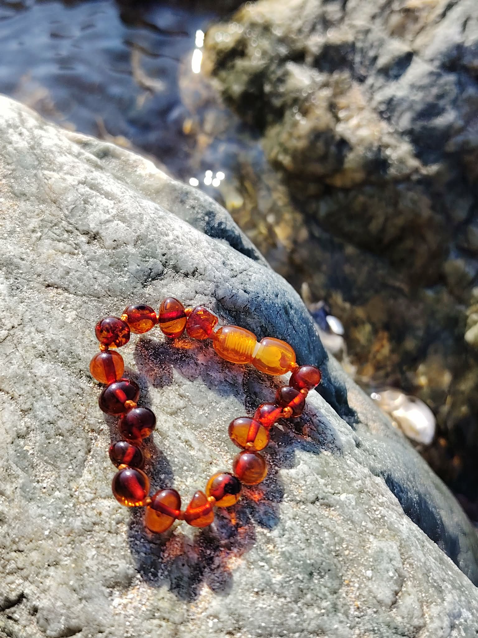 Bracelet en ambre pour bébé posé sur galet près de l’eau, fermoir de sécurité et nuances dorées, accessoire tendre au style naturel.