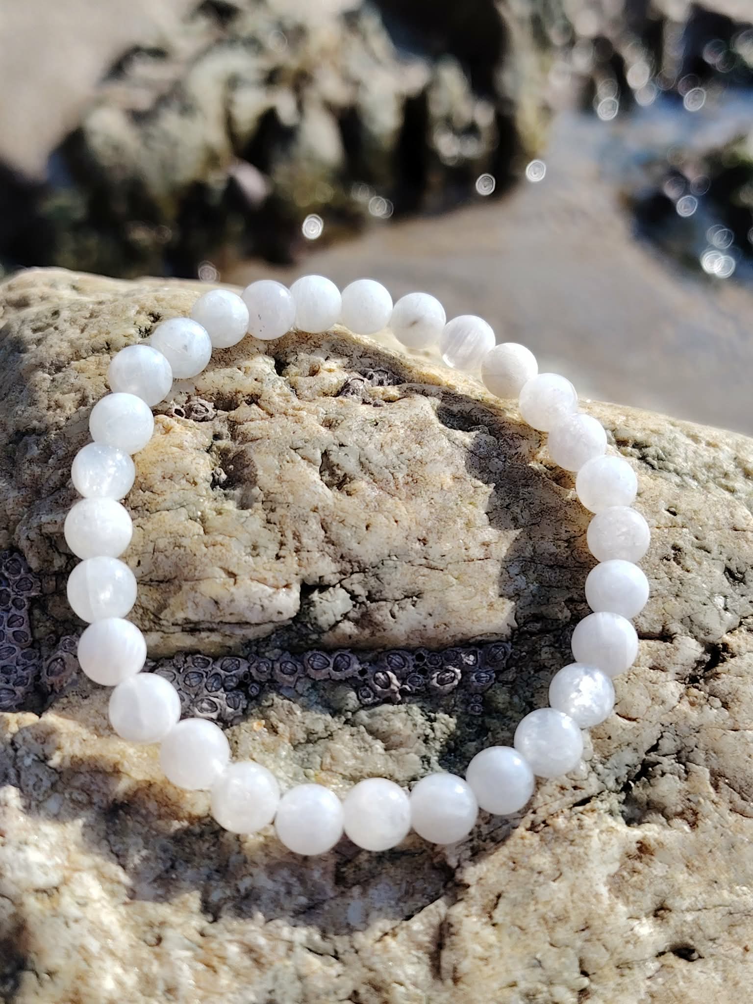 Bracelet en pierre de lune 6 mm posé sur les rochers en Bretagne près de l’eau, dans une ambiance douce et minérale évoquant intuition, sérénité