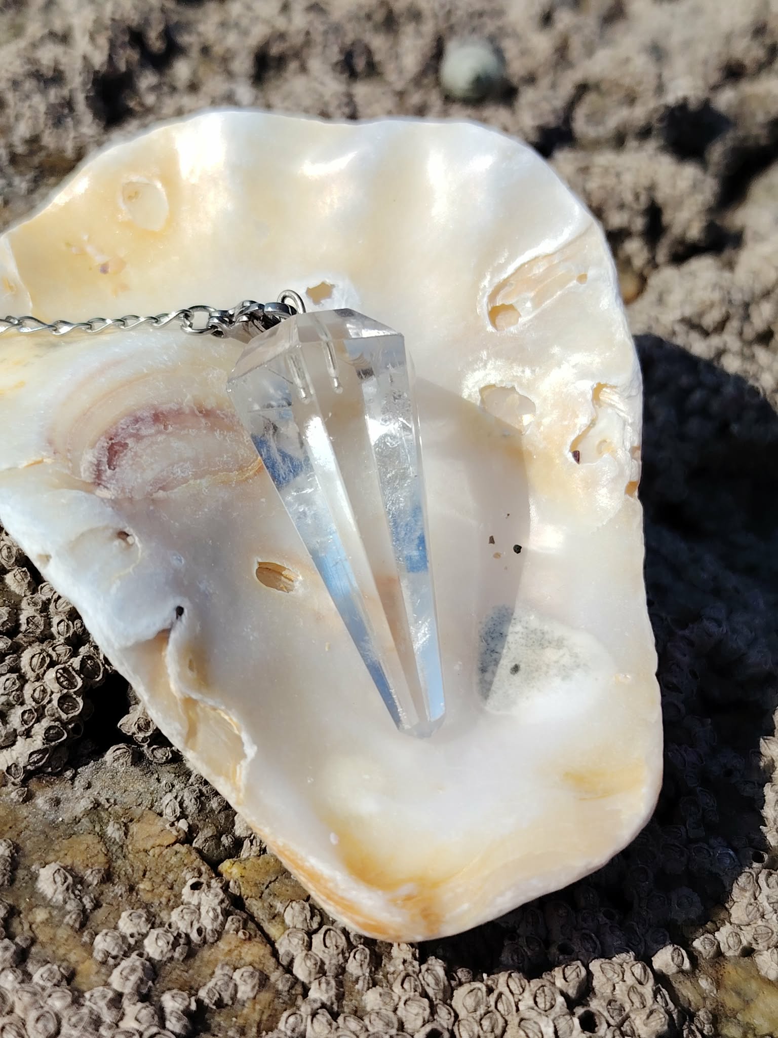 Pendule en cristal de roche posé dans un coquillage sur les rochers en Bretagne, dans une ambiance minérale lumineuse évoquant clarté, intuition et recentrage.