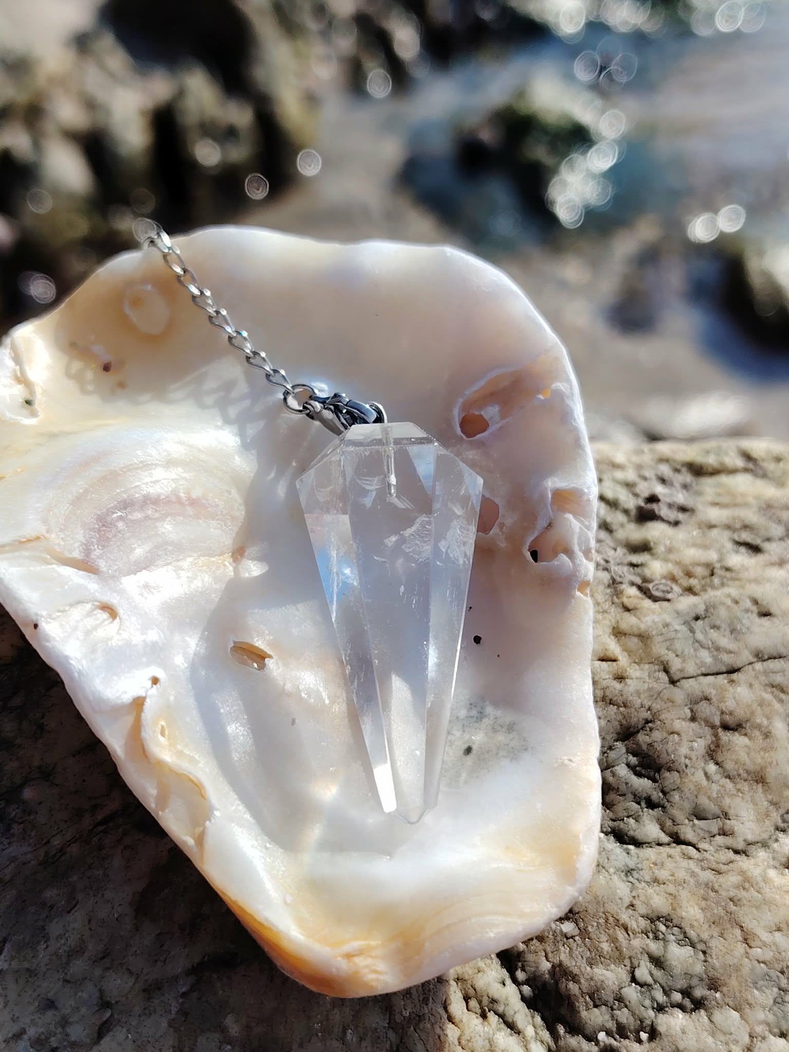 Pendule en cristal de roche présenté dans un coquillage sur la côte bretonne, entre roche et reflets marins, dans une atmosphère pure évoquant clarté, guidance et équilibre intérieur.