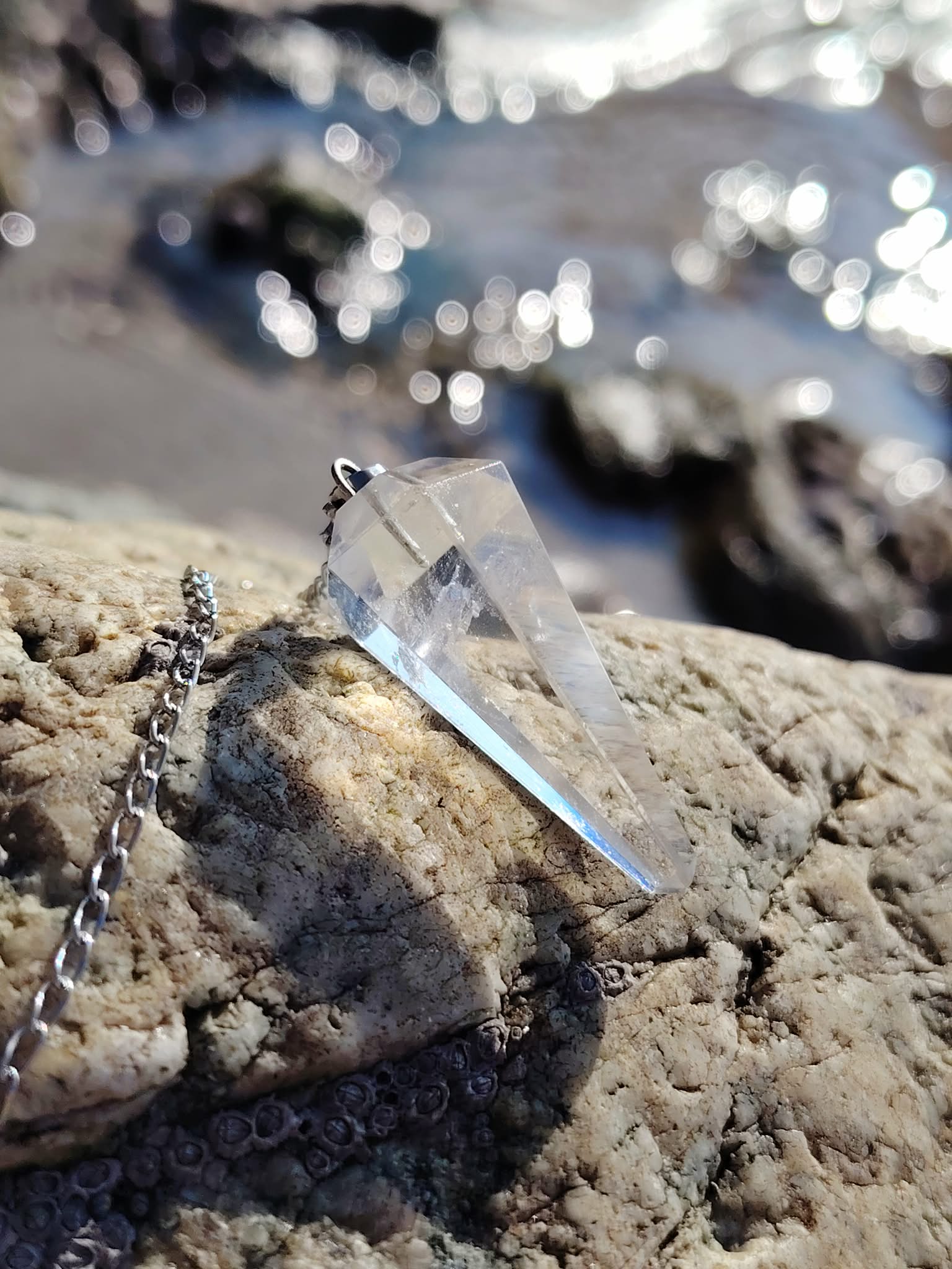 Pendule en cristal de roche personnalisable ou non, posé sur les rochers en Bretagne face aux reflets de l’eau, dans une ambiance lumineuse évoquant clarté, recentrage et intuition