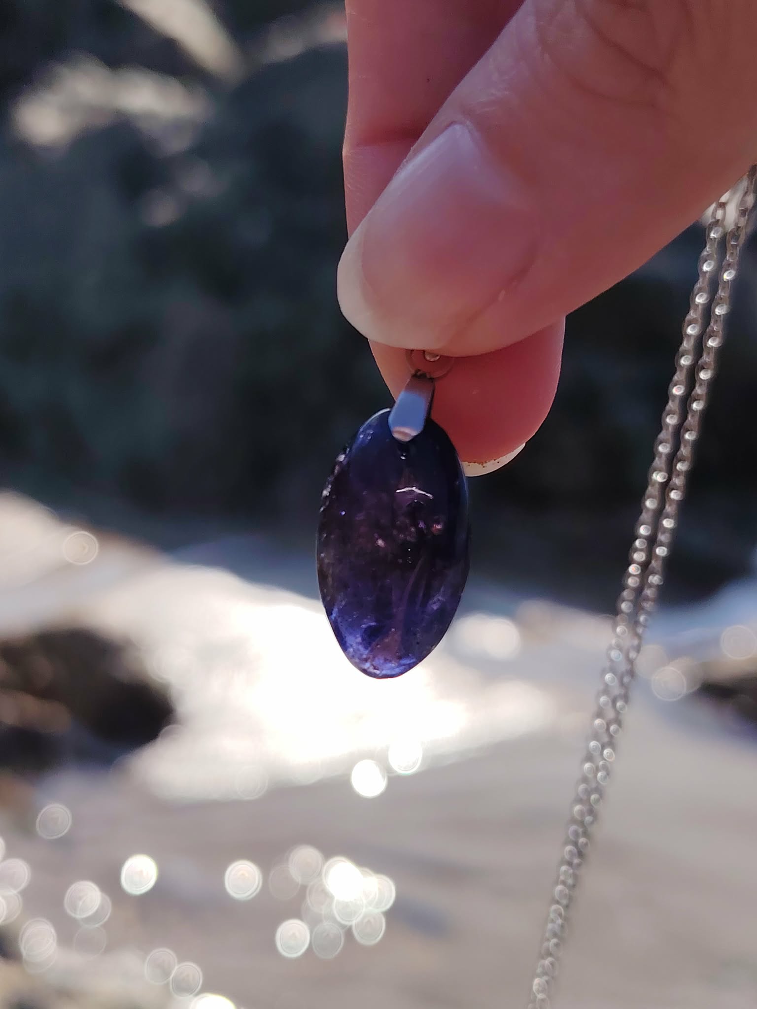 Main tenant un pendentif en iolite ou cordiérite au-dessus de l’eau en Bretagne, bijou en pierre naturelle associé à la fluidité intérieure, au recentrage et à la sérénité.