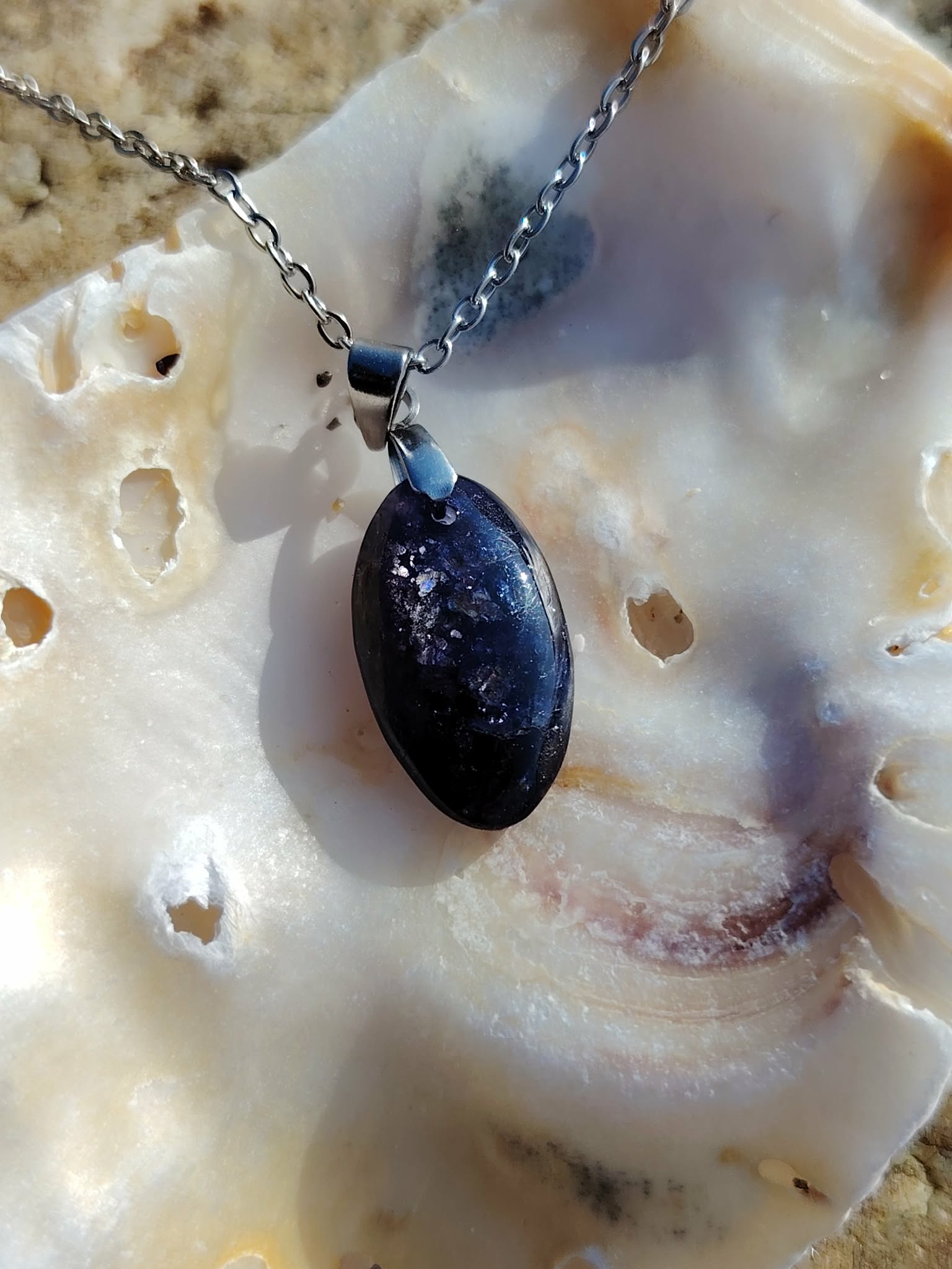 Pendentif en iolite ou cordiérite sur chaîne argentée, présenté dans un coquillage sur rochers en Bretagne, bijou en pierre naturelle évoquant la douceur, l’introspection et l’harmonie intérieure.