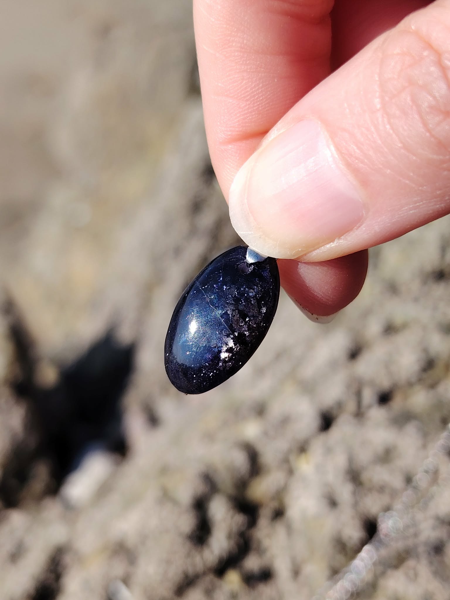 Main tenant un pendentif en iolite ou cordiérite devant les rochers en Bretagne, bijou en pierre naturelle évoquant le discernement, la confiance douce et la paix du mental.