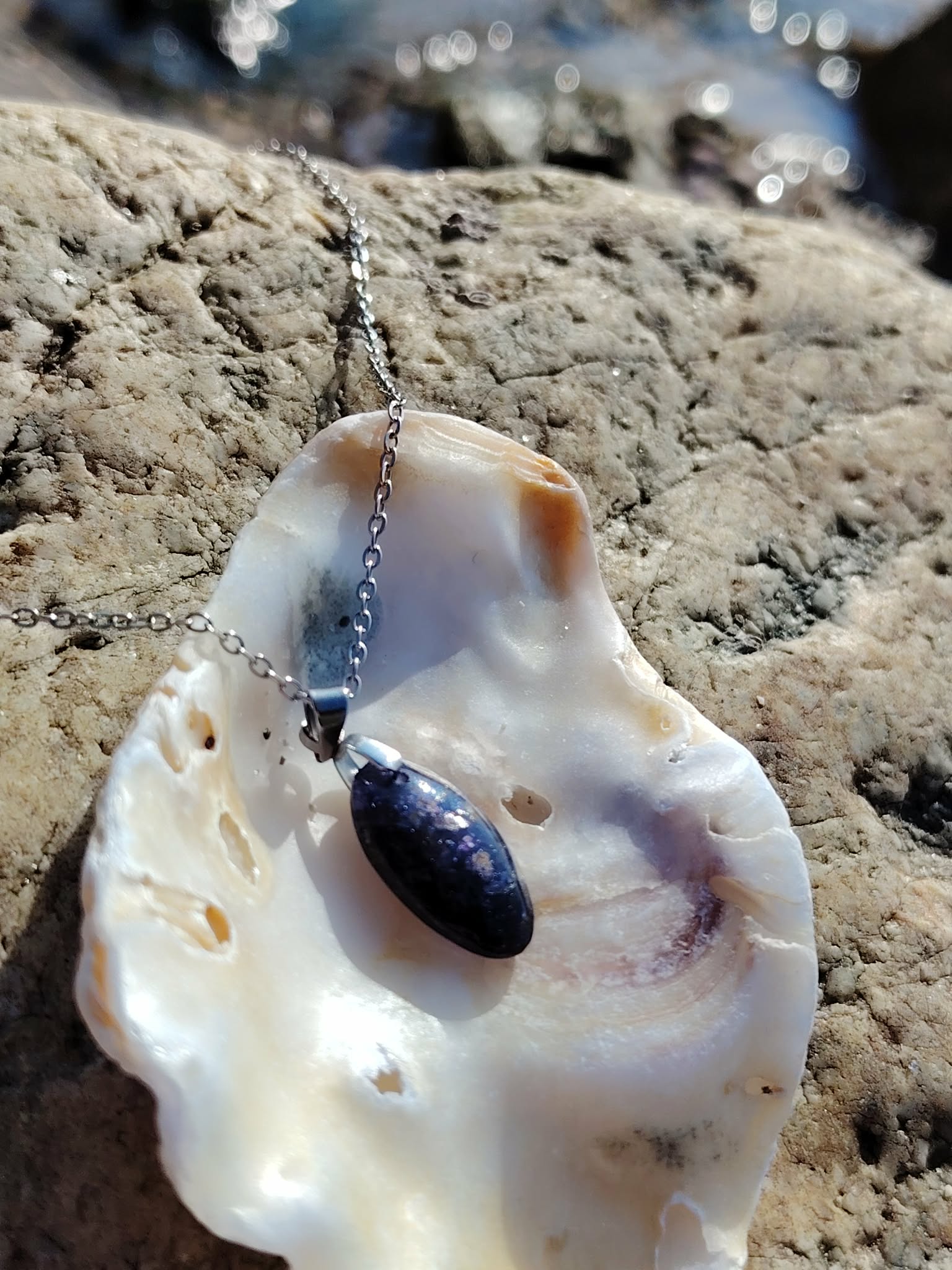 Pendentif en iolite ou cordiérite sur chaîne argentée, présenté sur un coquillage posé sur rochers en Bretagne, pierre naturelle reliée à la sagesse intérieure, au calme émotionnel et à la finesse intuitive.