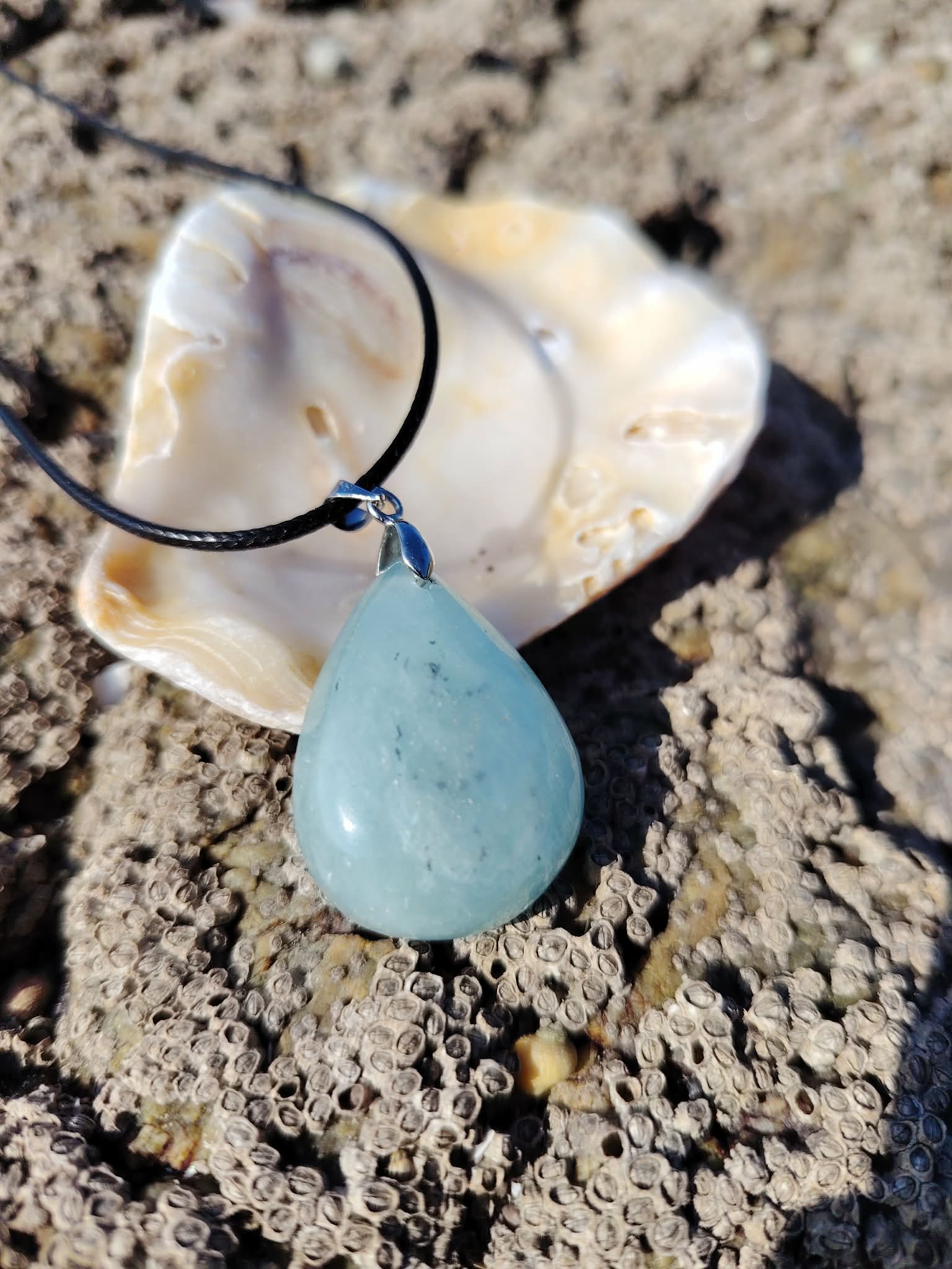 Pendentif en aigue-marine sur cordon noir avec coquillage sur rochers en Bretagne, pierre naturelle reliée à la paix du mental, à la fluidité intérieure et à l’écoute de soi.