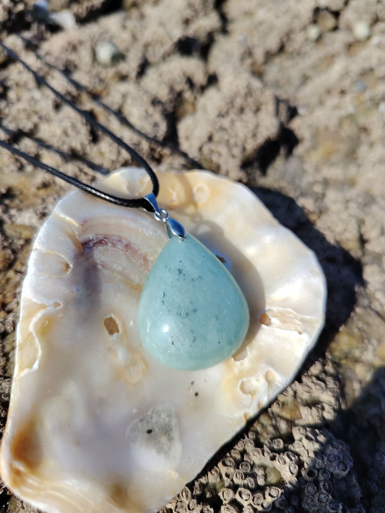 Pendentif en aigue-marine sur cordon noir, présenté dans un coquillage sur rochers en Bretagne, pierre naturelle associée à la sérénité, à la clarté émotionnelle et à la douceur océane.