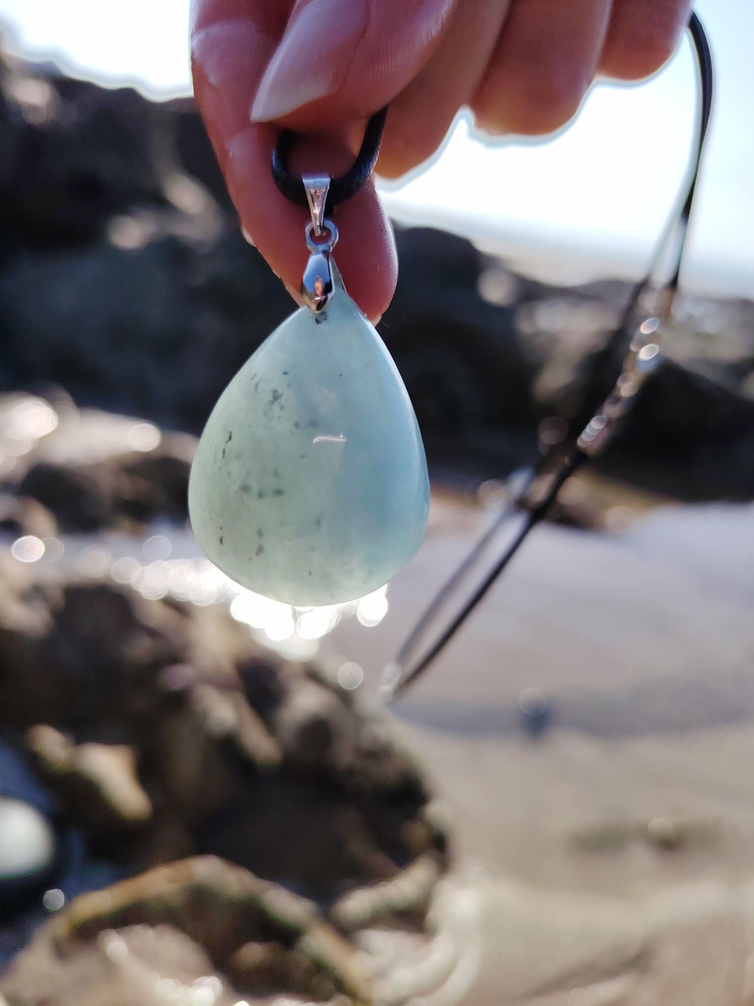 Main tenant un pendentif en aigue-marine au-dessus des rochers et de l’eau en Bretagne, bijou en pierre naturelle lié à la fluidité, à la sensibilité paisible et au recentrage.