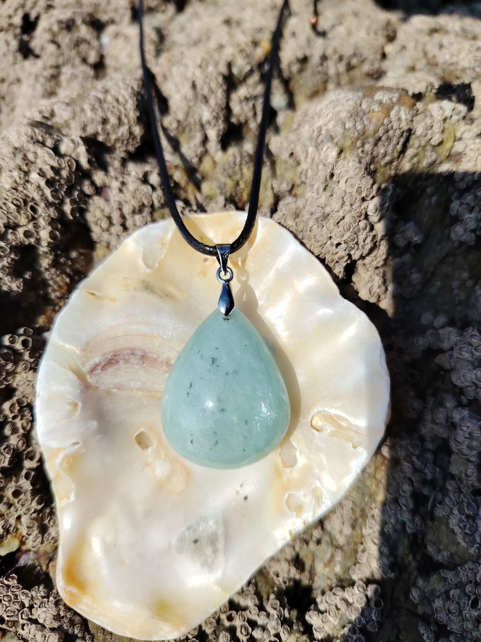 Pendentif en aigue-marine sur cordon noir, présenté dans un coquillage posé sur rochers en Bretagne, pierre naturelle évoquant la douceur marine, l’apaisement et la respiration intérieure.