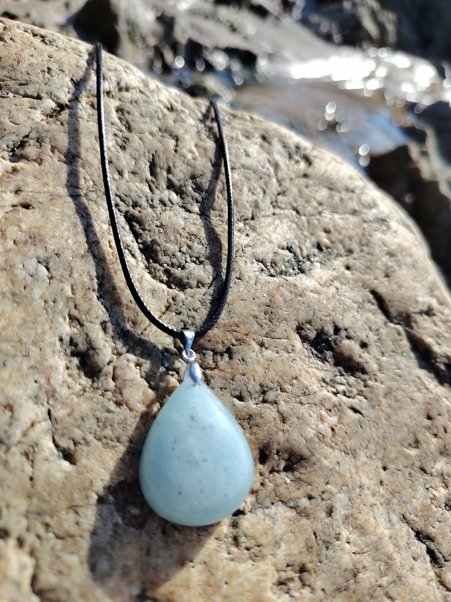 Pendentif en aigue-marine sur cordon noir, posé sur rochers en Bretagne près de l’eau, bijou en pierre naturelle associé à la sérénité, à la fluidité émotionnelle et à la paix intérieure.