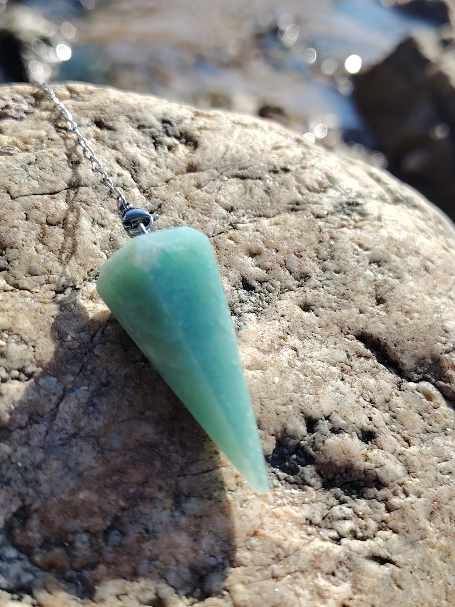 Pendule en amazonite vert d’eau posé sur un rocher en Bretagne, avec fond marin flou, évoquant douceur, calme intérieur, clarté et harmonie.