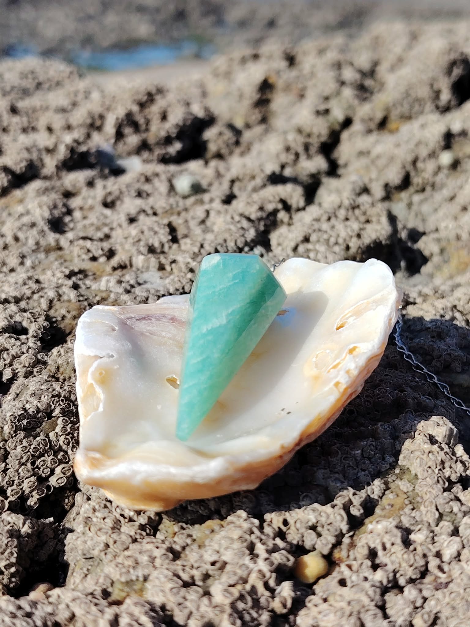 Pendule en amazonite bleu-vert présenté dans un coquillage sur roche en Bretagne, image douce inspirant calme, intuition et harmonie.