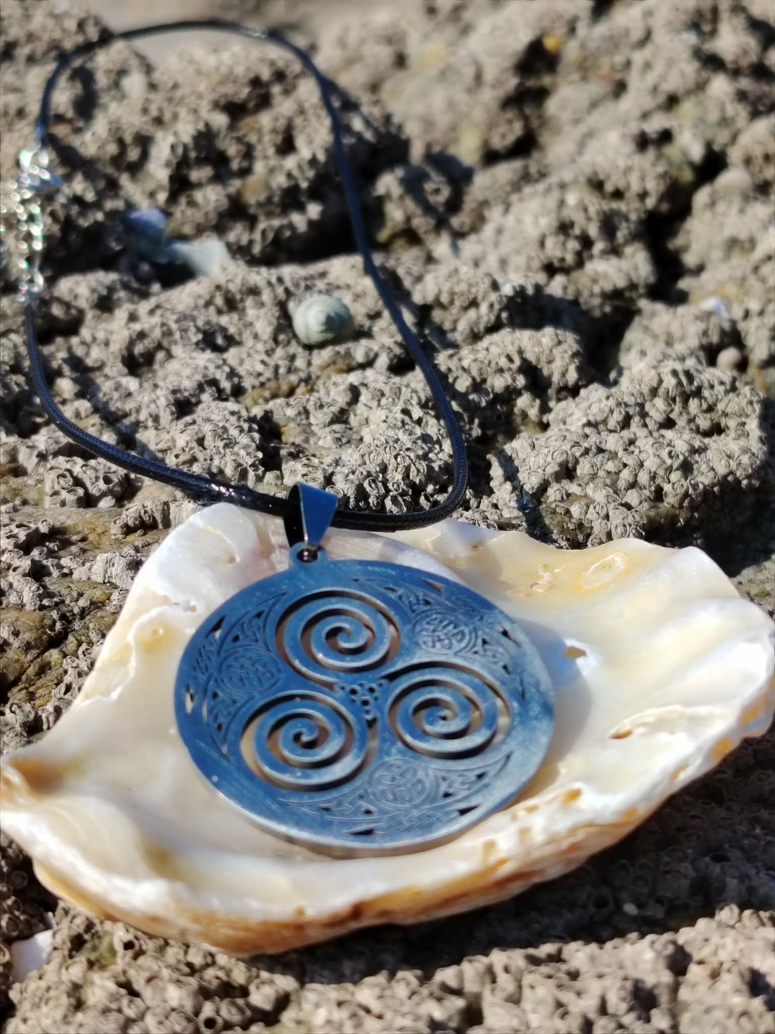 Pendentif Triskell sur cordon noir présenté sur un coquillage parmi les rochers en Bretagne, symbole d’évolution et d’harmonie