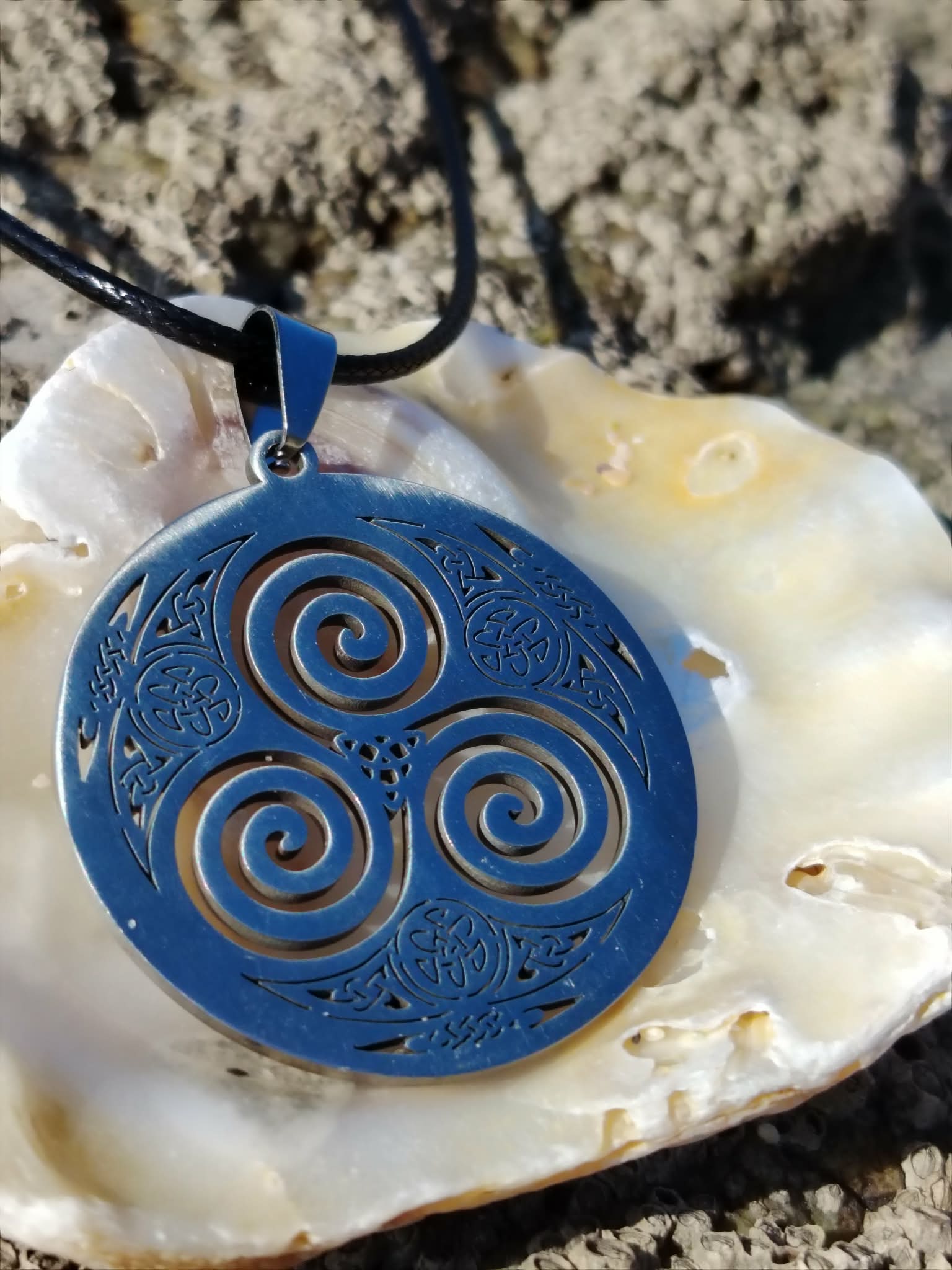 Pendentif Triskell bleu sur cordon noir présenté sur un coquillage parmi les rochers en Bretagne, bijou celtique au style minéral