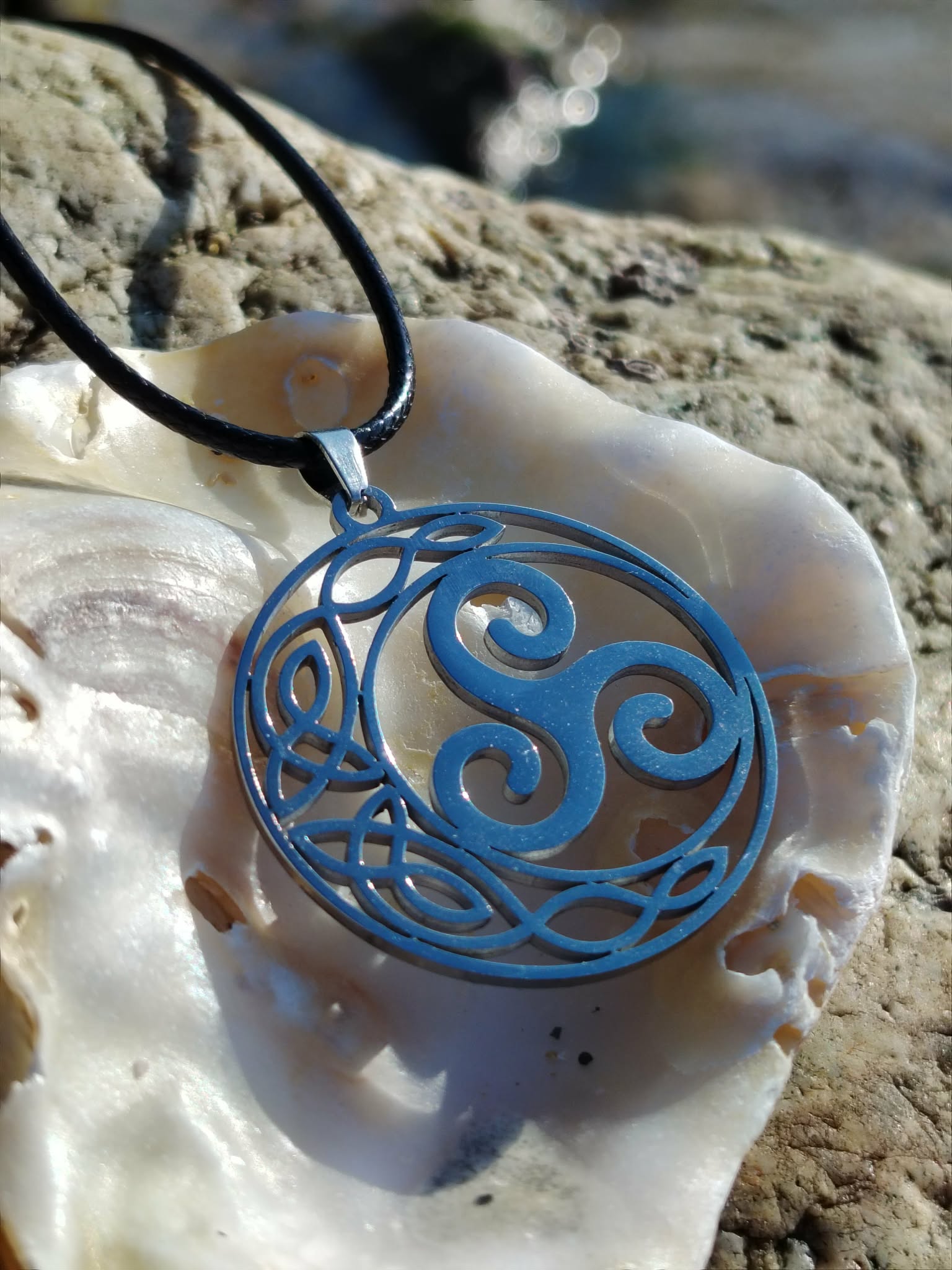Pendentif Triskell bleu sur coquillage en décor breton minéral, suspendu à un cordon noir, symbole d’unité et d’énergie cyclique