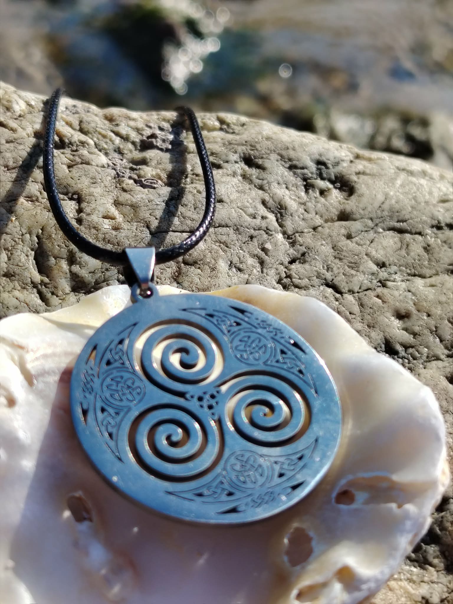 Pendentif Triskell bleu dans un coquillage sur rochers bretons, cordon noir et symbole celtique de vie en mouvement