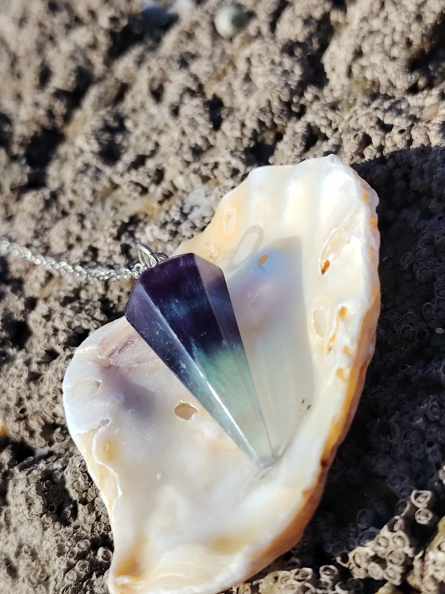 Pendule en fluorite posé dans un coquillage sur un décor minéral, outil de divination à l’ambiance douce et mystique évoquant intuition, clarté mentale et recentrage.