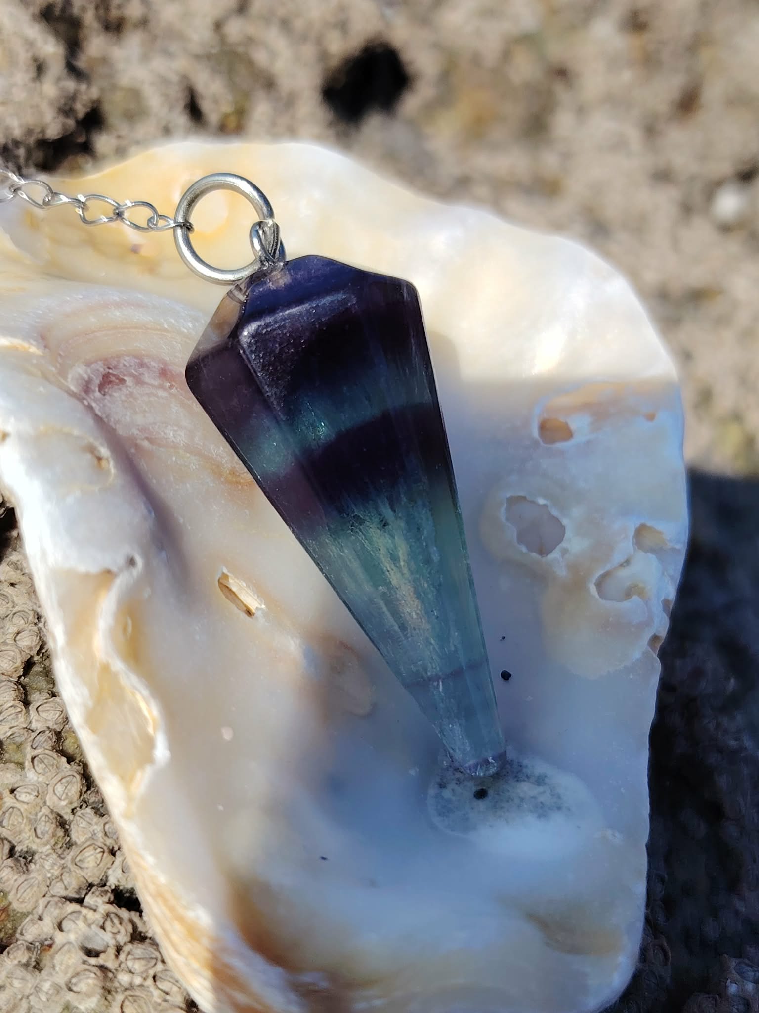 Pendule en fluorite posé dans un coquillage clair sur fond sableux, outil de divination à l’ambiance douce évoquant intuition, clarté mentale et recentrage.