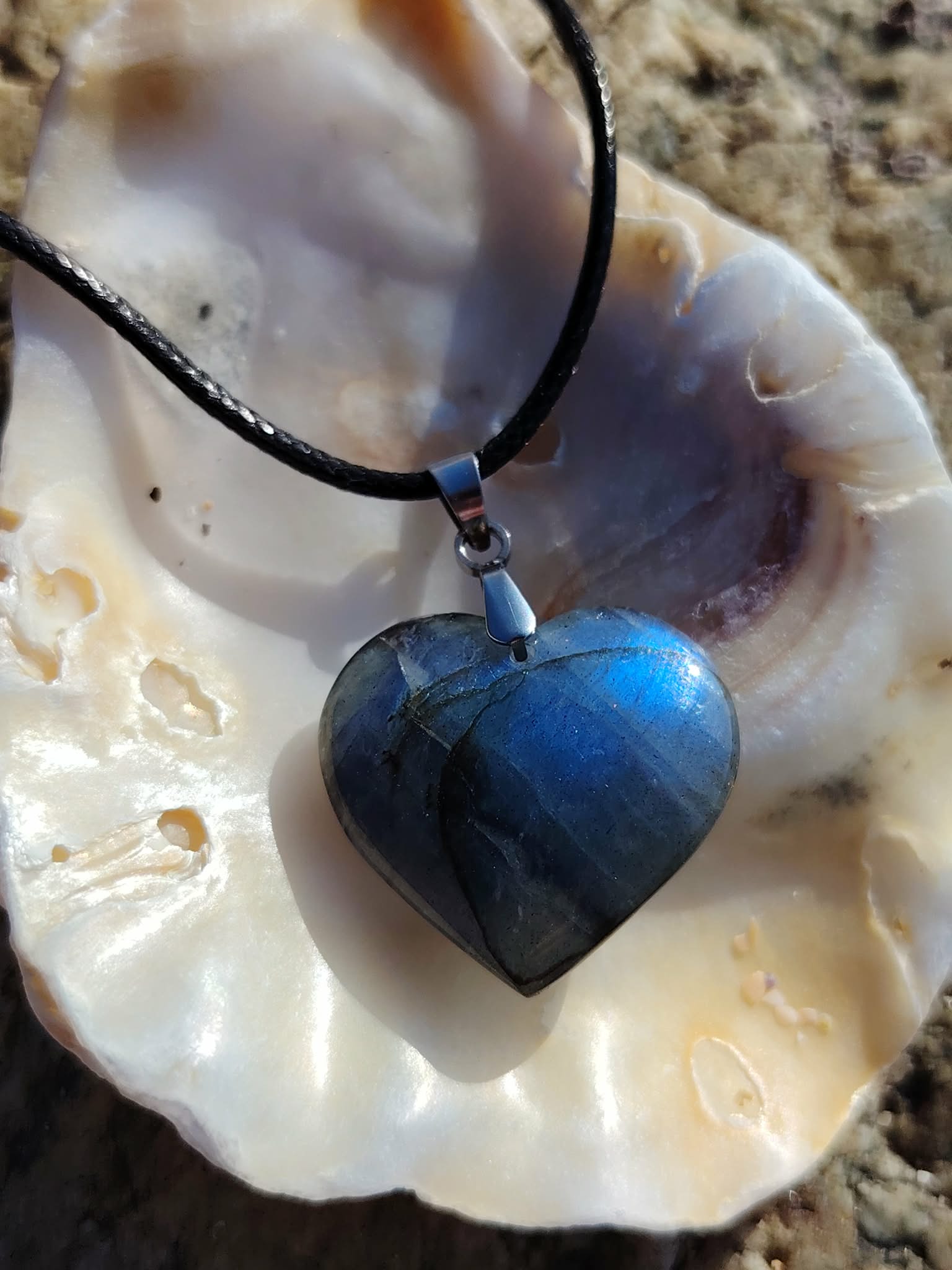 Pendentif cœur en labradorite sur cordon noir, présenté dans un coquillage en Bretagne, collier pierre naturelle relié à la sensibilité, au calme intérieur et à la perception fine.